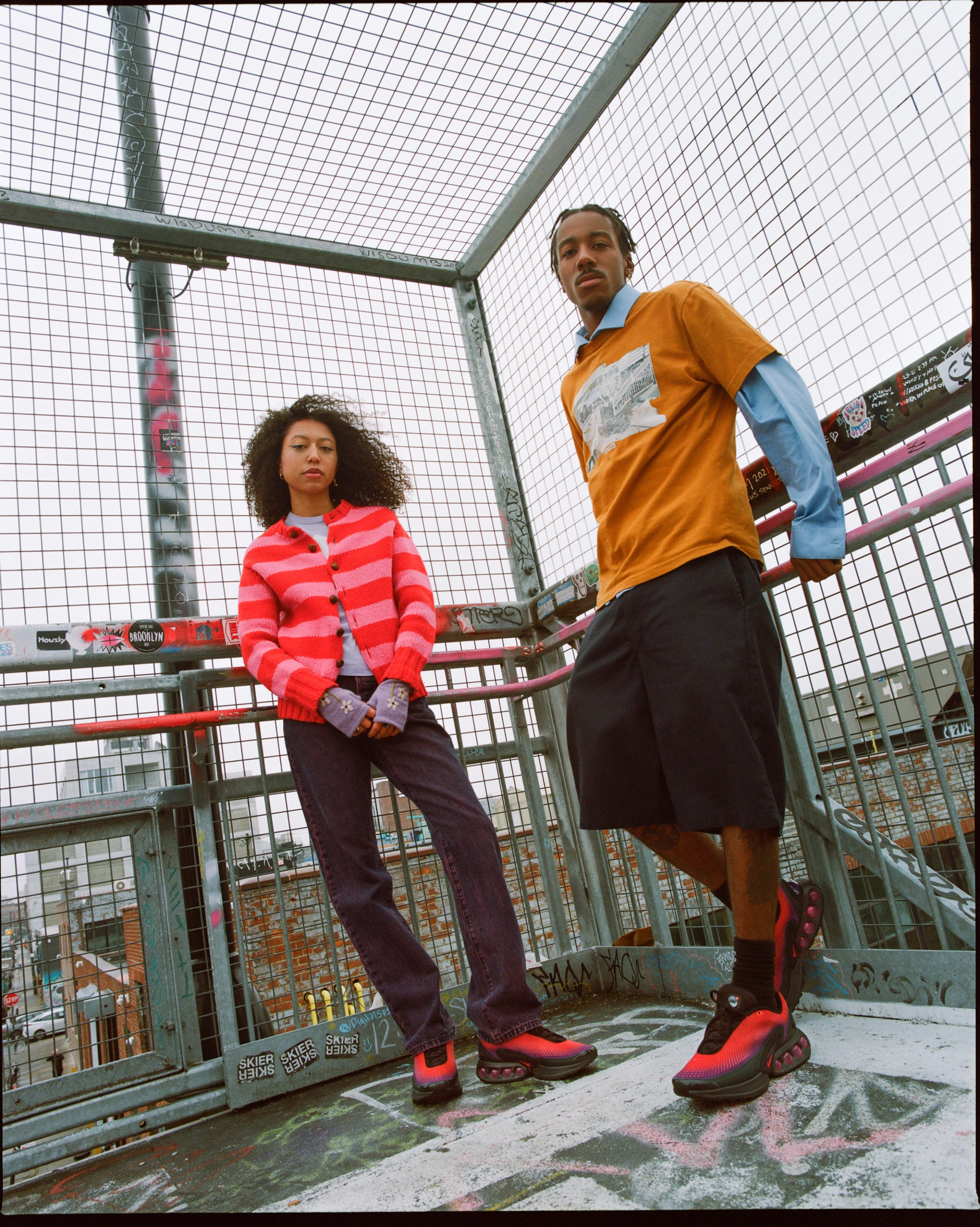 A male and female model both wearing the Nike Air Max Dn in a black and red color while standing outside against a fenced off railing.