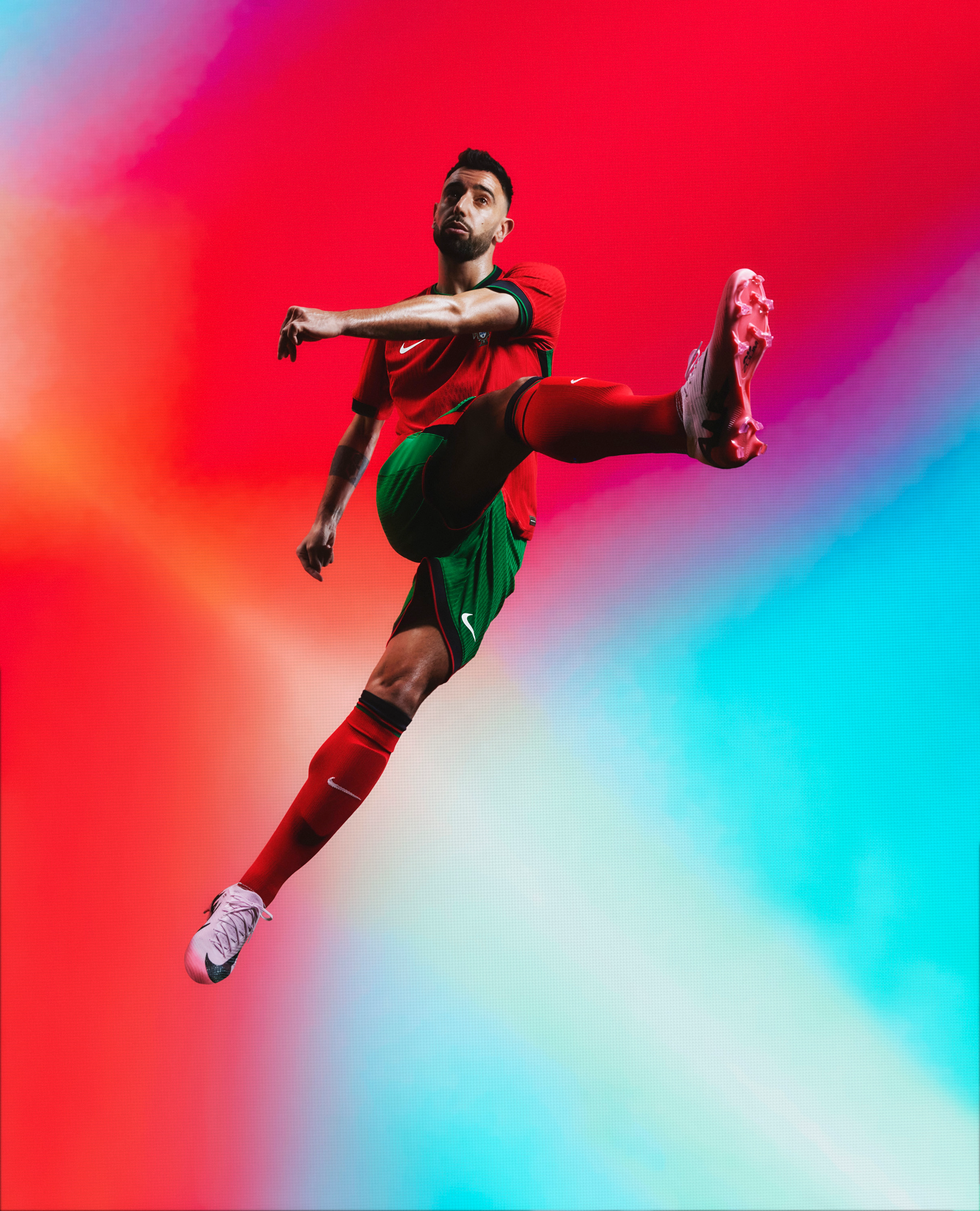 Athlete in a kicking pose wearing a red jersey with a forest green collar and forest green cuffs, as well as matching forest green shorts with red trim with a Nike swoosh and team logo on the chest and legs. They are also wearing matching red socks with green trim.