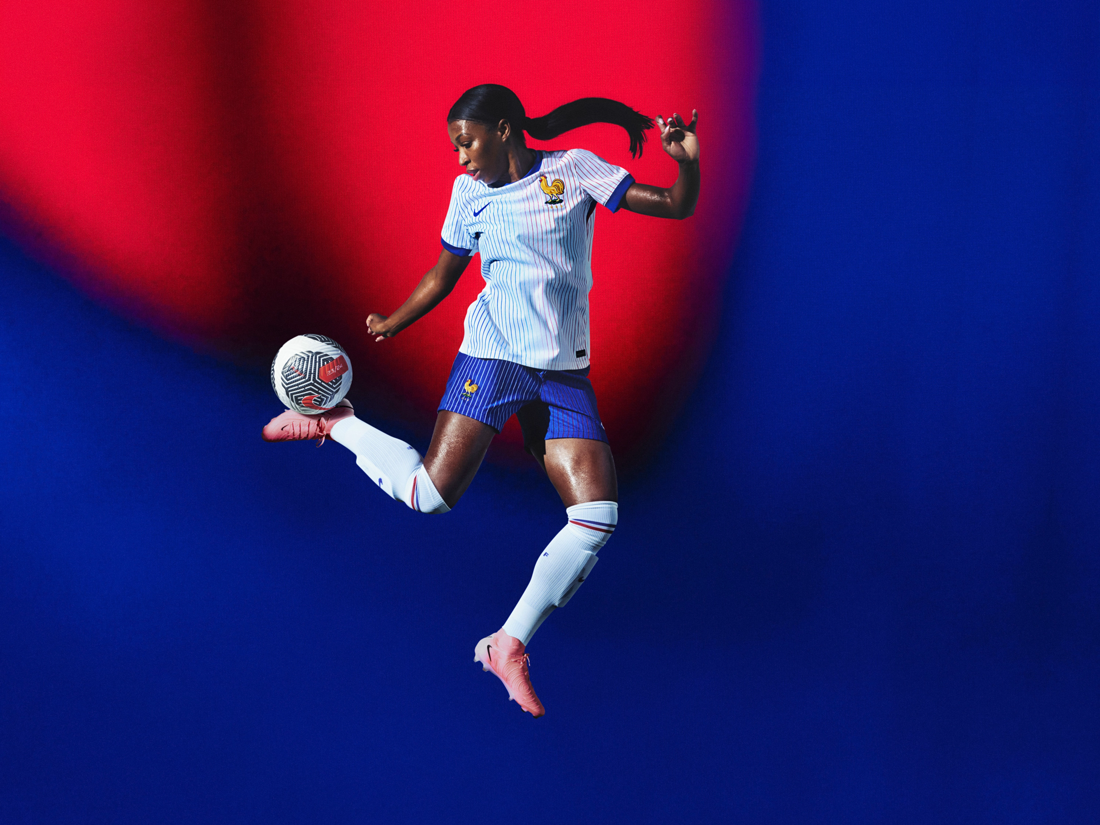 Athlete bouncing a soccer ball off their heel wearing a white soccer jersey with red and blue pinstripes, as well as cobalt blue shorts with white pinstripes, each with a Nike swoosh and team logo on the chest and leg. They are also wearing white socks with red and blue striped trim.