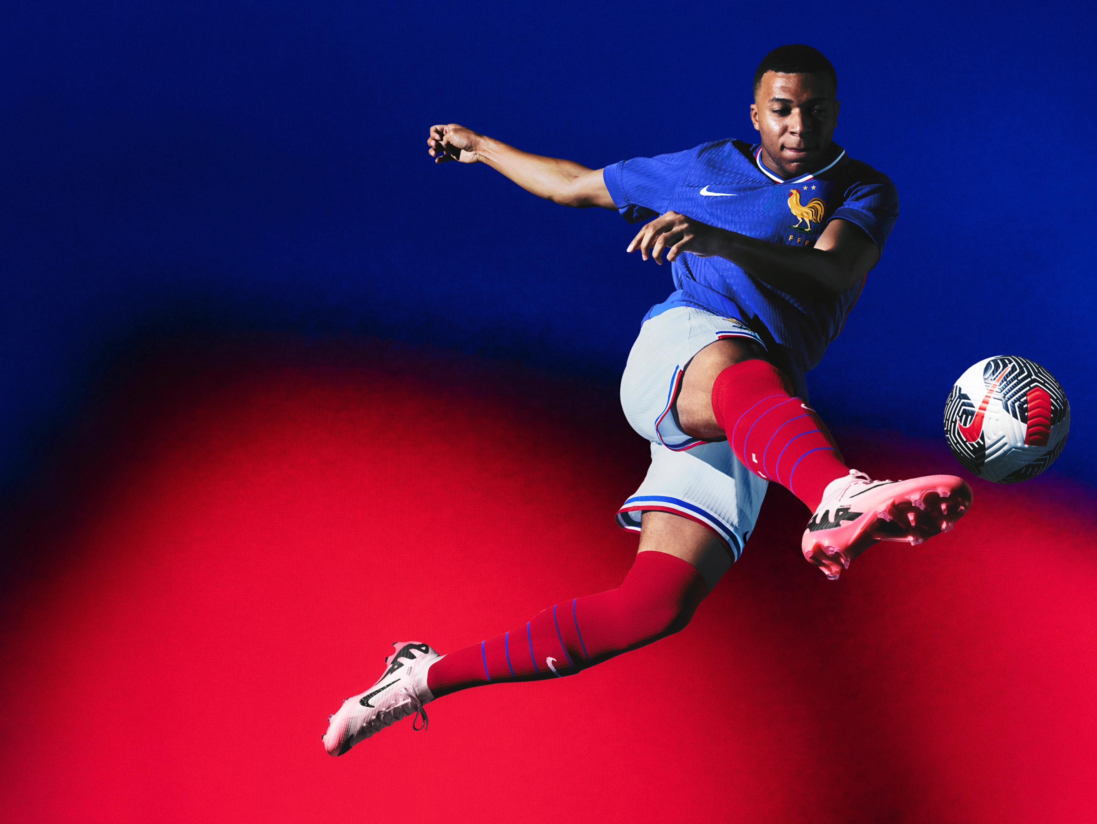 Athlete in a flying kicking pose wearing a cobalt blue soccer jersey, as well as white shorts with blue and red striped trim, each with a Nike swoosh and team logo on the chest and legs. They are also wearing red socks with blue horizontal stripes on the legs.