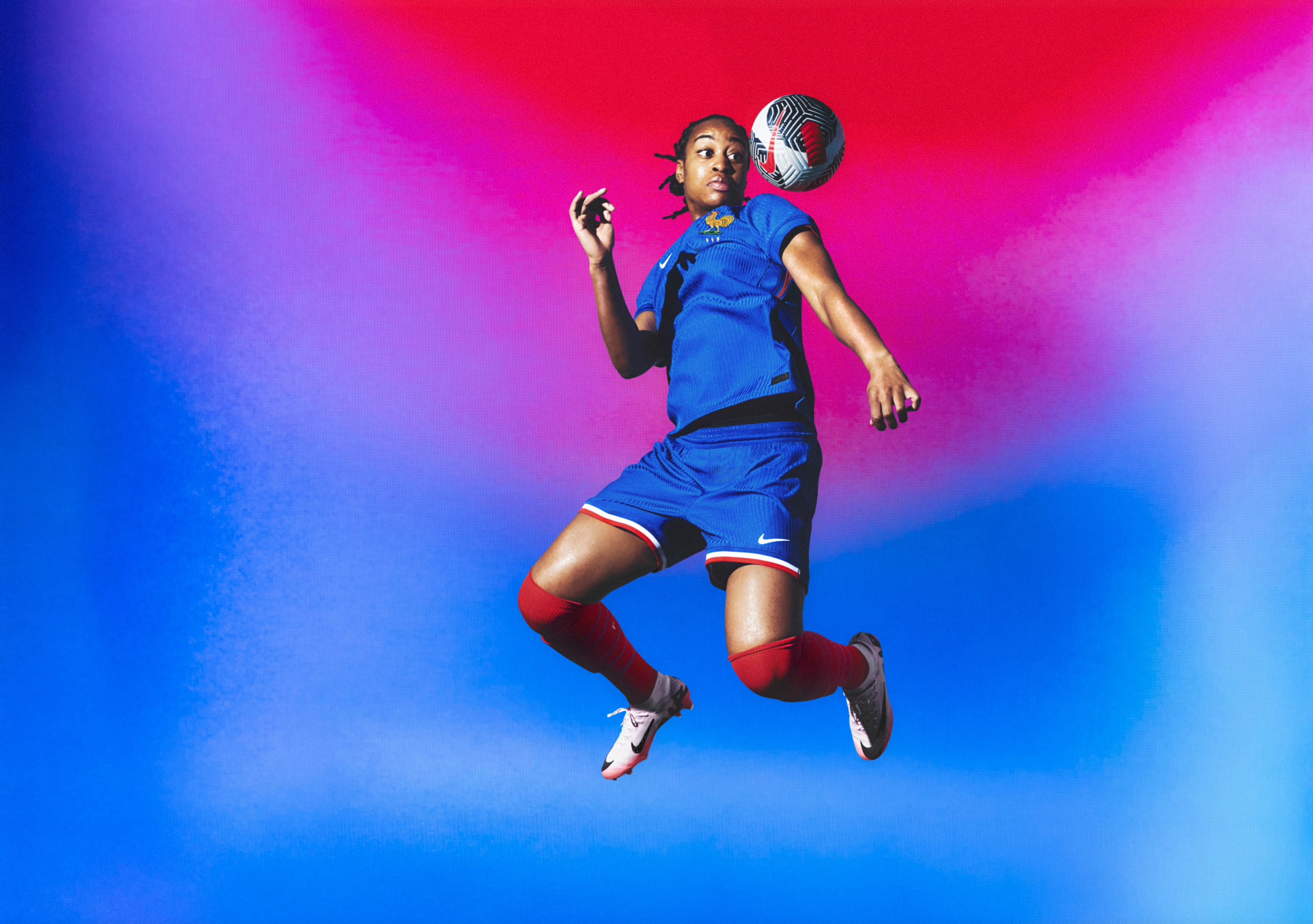 Athlete bouncing a soccer ball off their shoulder wearing a cobalt blue soccer jersey, as well as matching cobalt blue shorts with red, white, and blue striped trim. They are also wearing red socks.