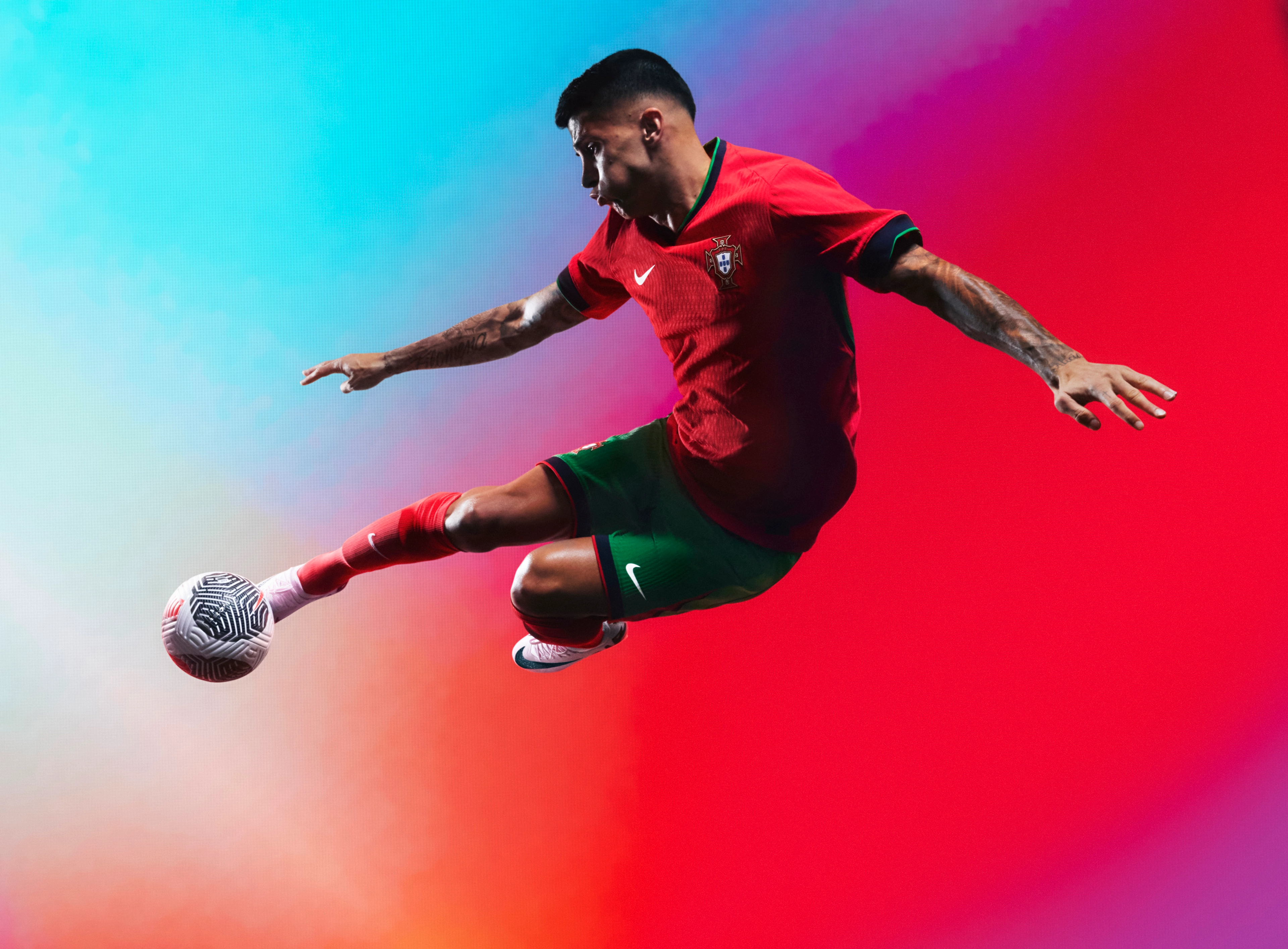 Athlete in a flying kicking pose wearing a red jersey with a forest green collar and forest green cuffs, as well as matching forest green shorts with red trim with a Nike swoosh and team logo on the chest and legs. They are also wearing matching red socks.