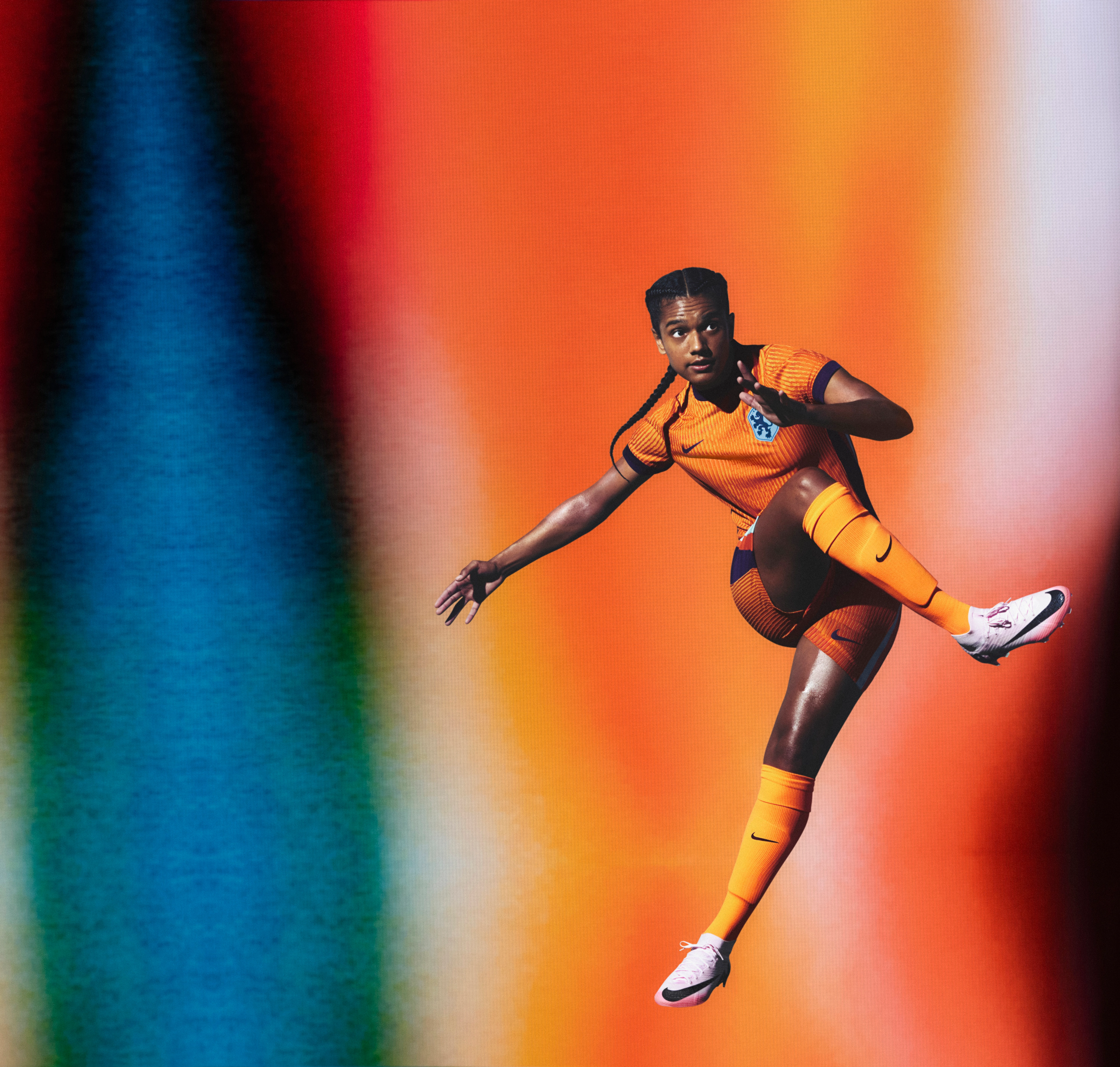 Athlete in a flying kicking pose wearing an orange soccer jersey and matching orange shorts with an indigo collar, cuffs, and sides, as well as matching shorts, each with a Nike swoosh and a team logo on the chest and legs. They are also wearing matching orange socks.
