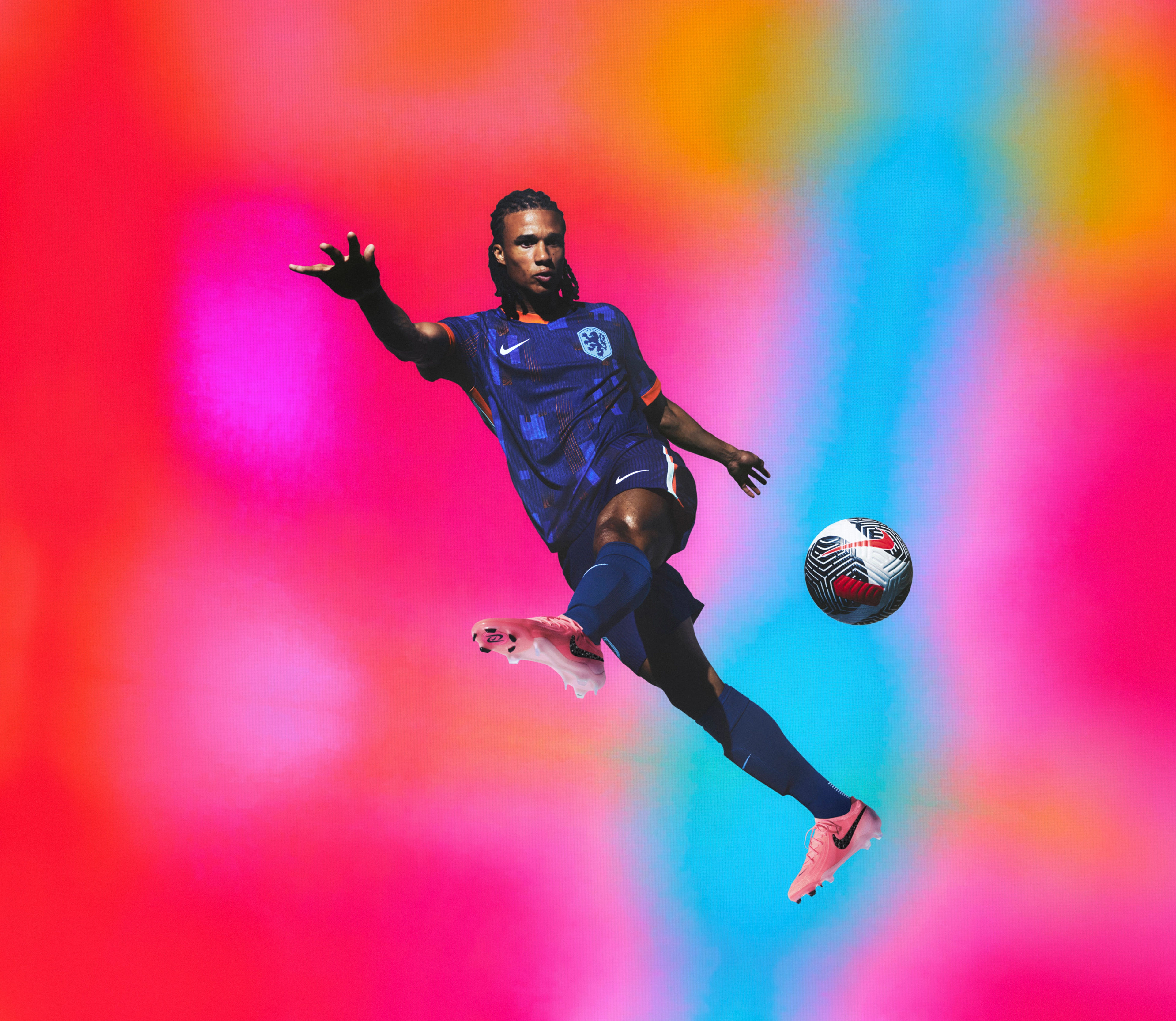 Athlete in a flying kicking pose wearing an indigo soccer jersey with an orange collar and cuffs and blue and orange rectangle patterns on the front, as well as matching indigo blue shorts, each with a Nike swoosh and a team logo on the chest and legs. They are also wearing indigo blue socks.