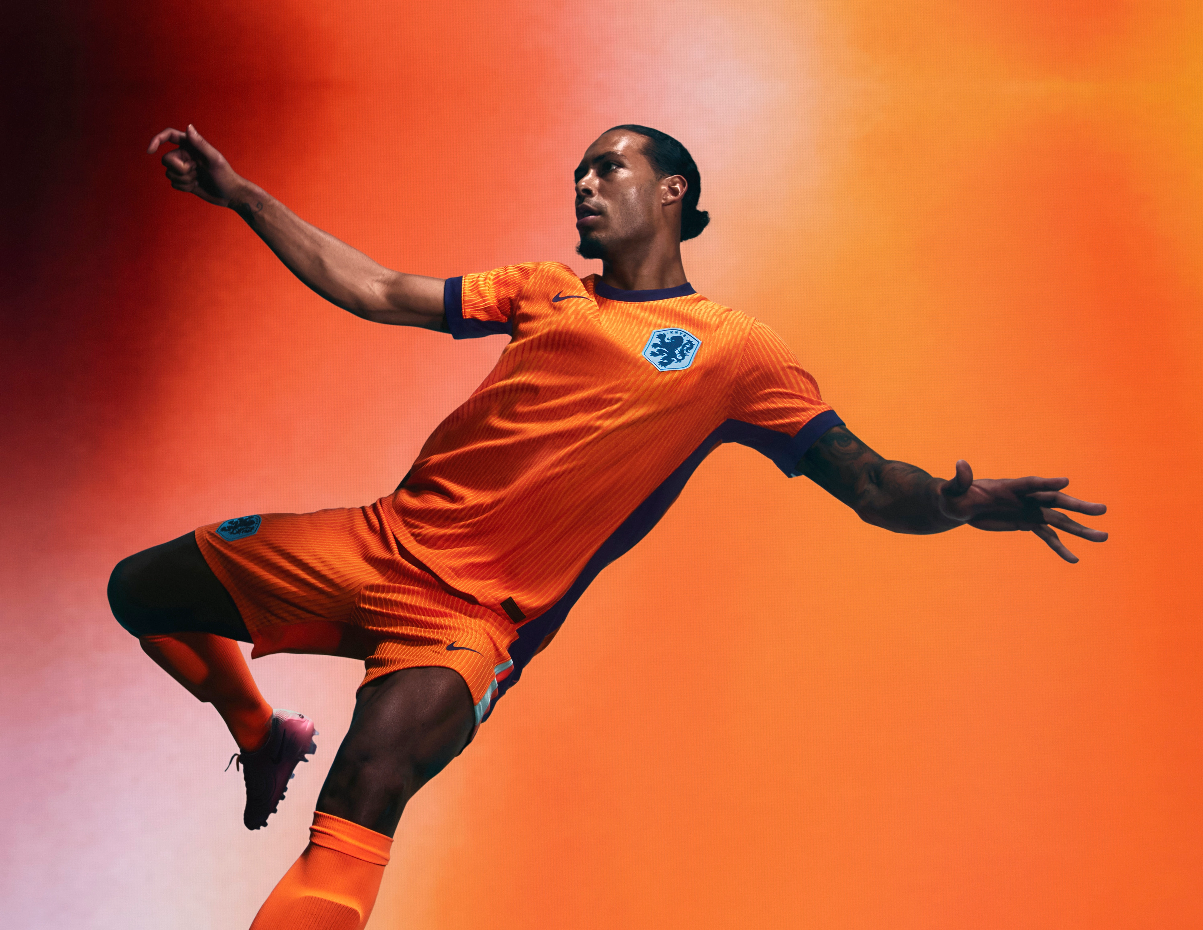 Athlete in a mid-kicking pose wearing an orange soccer jersey and matching orange shorts with an indigo collar, cuffs, and sides, as well as matching shorts, each with a Nike swoosh and a team logo on the chest and legs. They are also wearing matching orange socks.