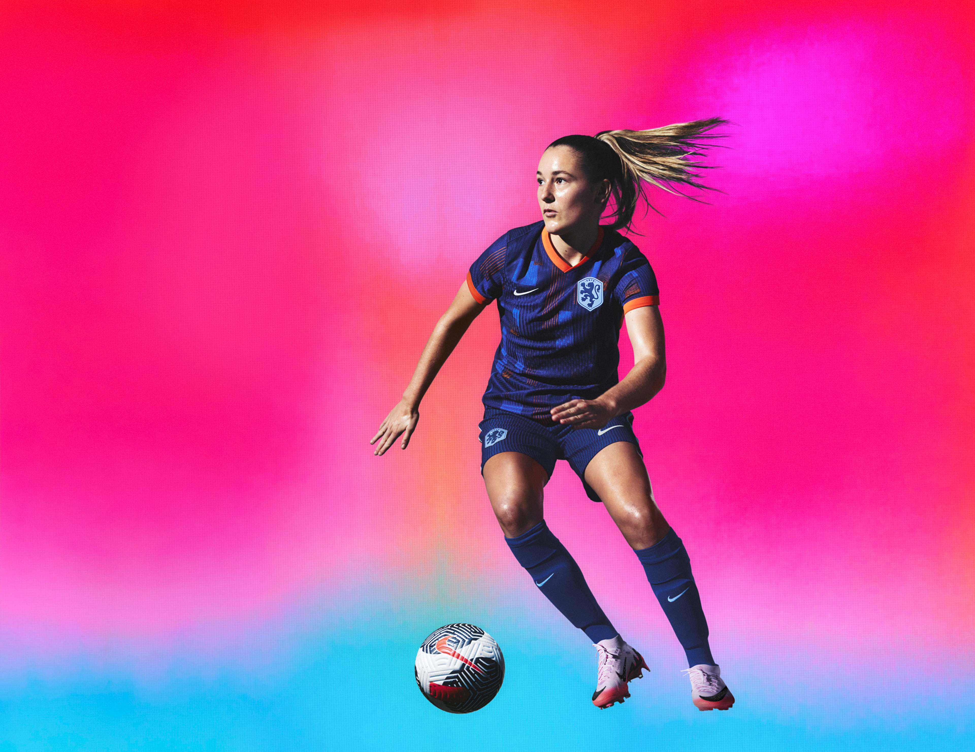 Athlete dribbling a soccer ball wearing an indigo soccer jersey with an orange collar and cuffs and blue and orange rectangle patterns on the front, as well as matching indigo blue shorts, each with a Nike swoosh and a team logo on the chest and legs. They are also wearing indigo blue socks.