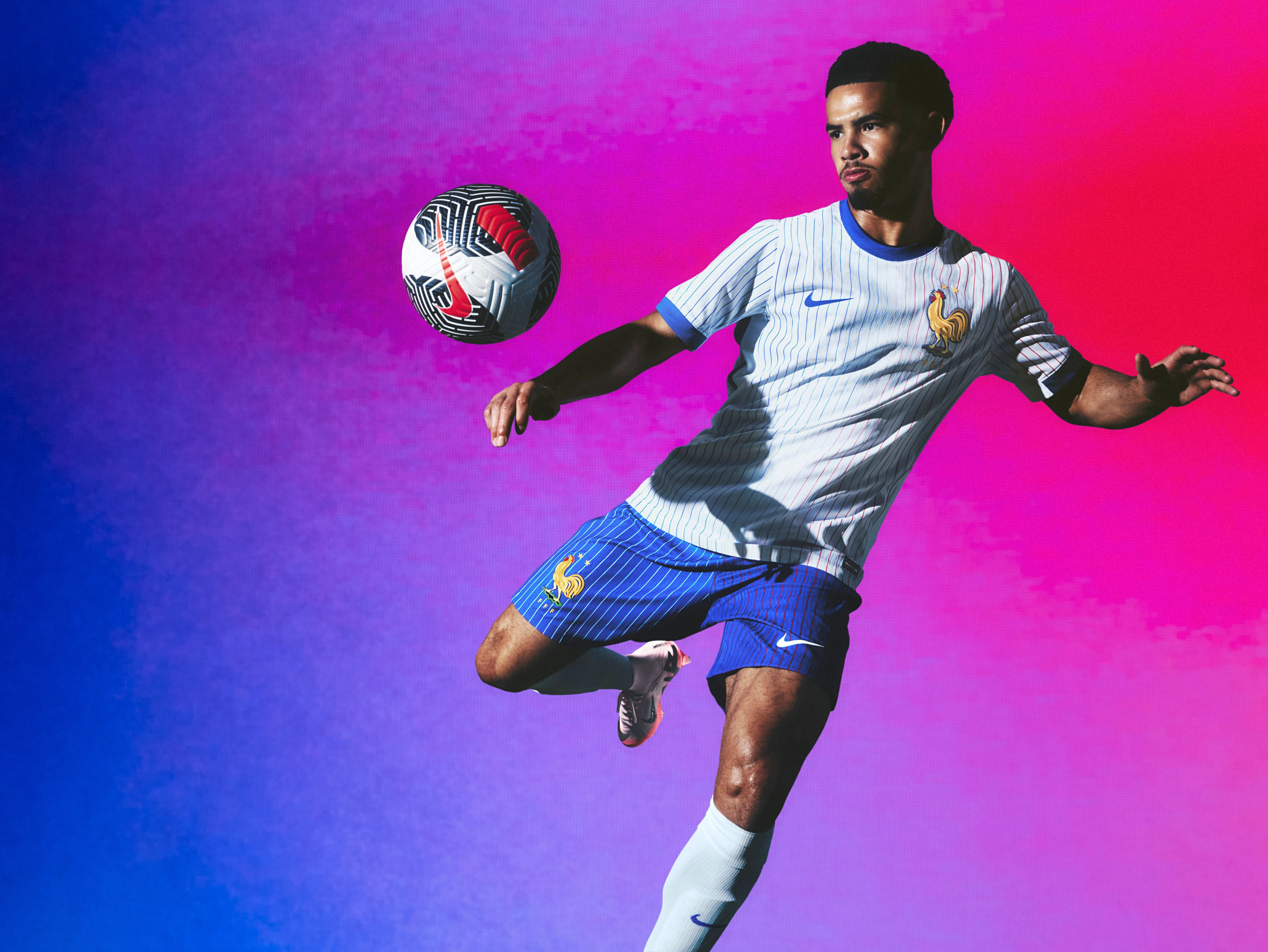 Athlete bouncing a soccer ball off their knee wearing a white soccer jersey with red and blue pinstripes, as well as cobalt blue shorts with white pinstripes, each with a Nike swoosh and team logo on the chest and leg. They are also wearing white socks.