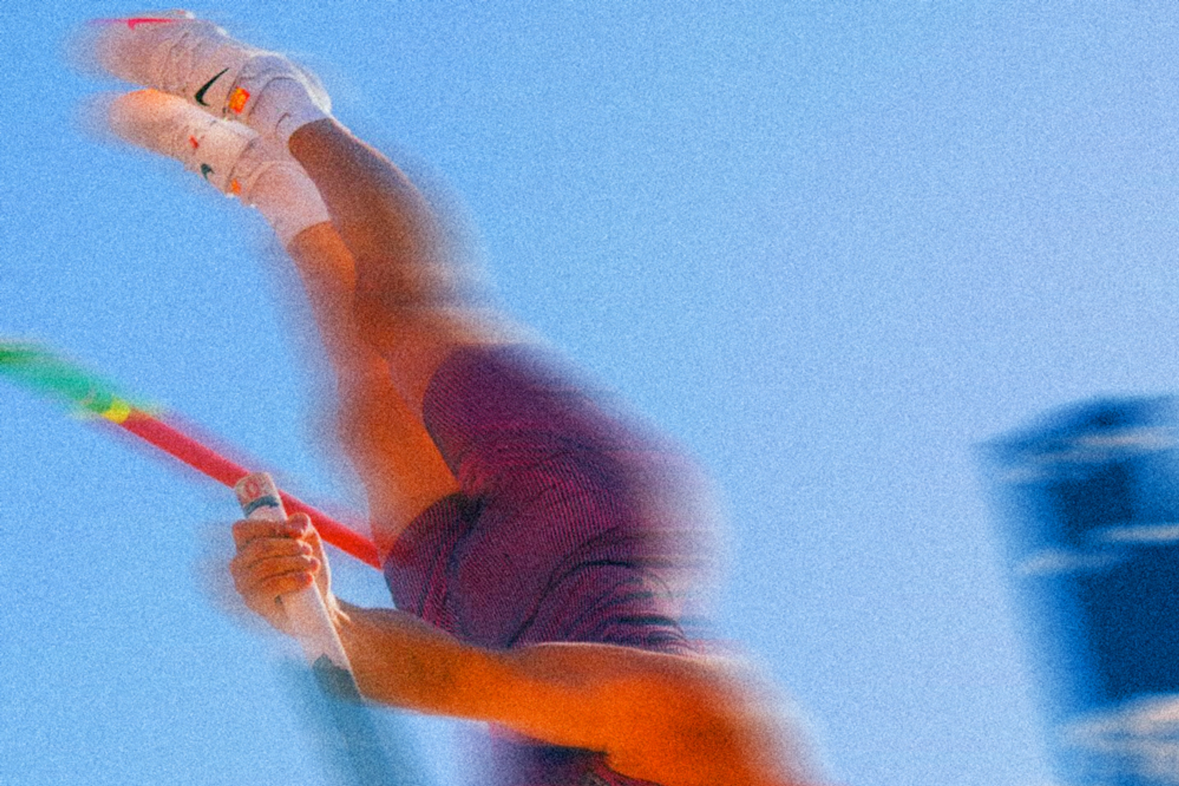 A blurred image of a pole vaulters legs as they clear the bar. You see the top of the pole, and white Nike shoes in the air, against a blue sky.