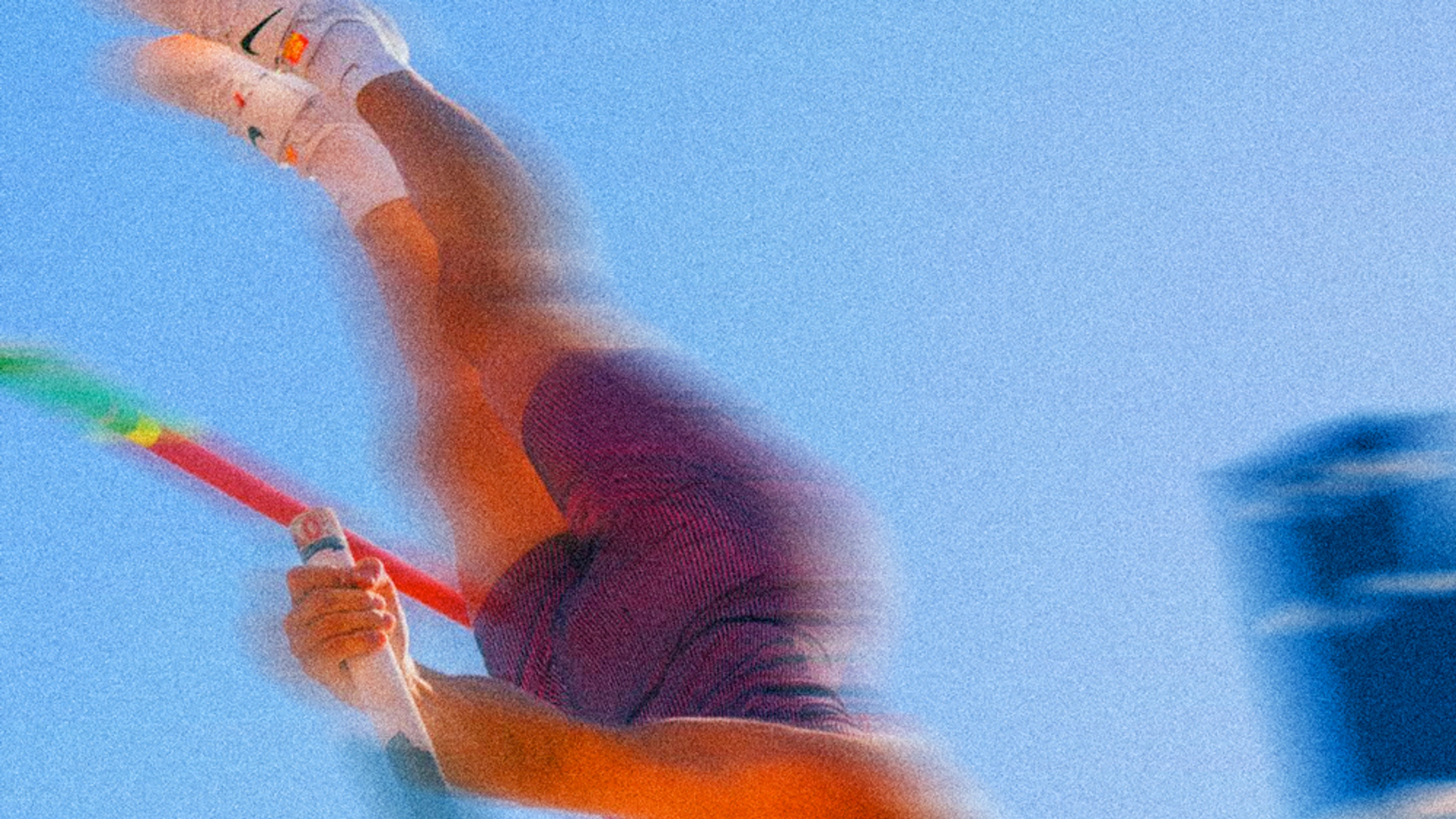 A blurred image of a pole vaulters legs as they clear the bar. You see the top of the pole, and white Nike shoes in the air, against a blue sky.