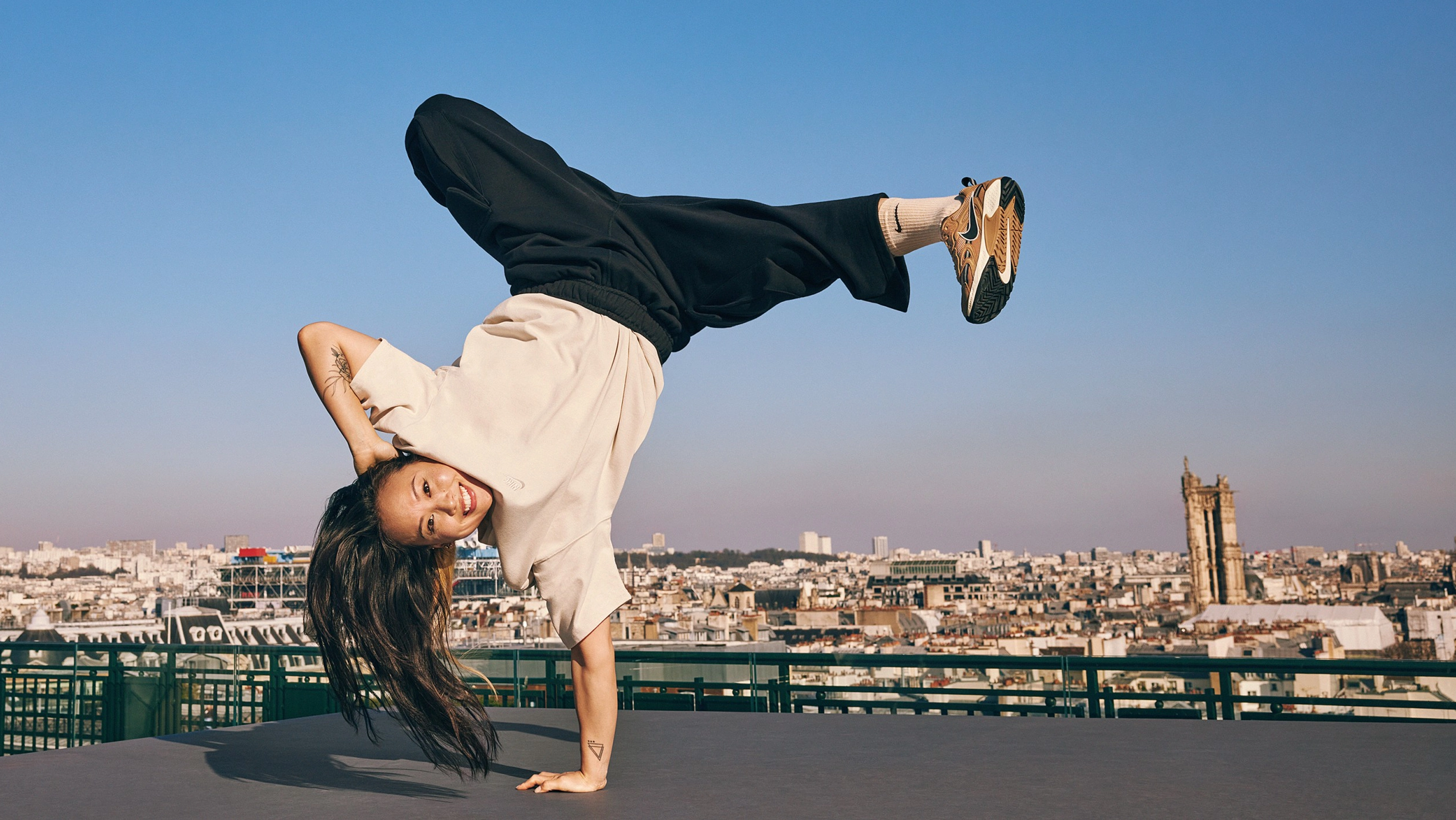 Sunny Choi does a one-handed handstand breakdancing move on a rooftop in Paris, France. Sunny wears black pants and a cream colored top from the Nike Sportswear collection, plus white Nike socks. The Paris city-scape with a bright blue sky is in the background.