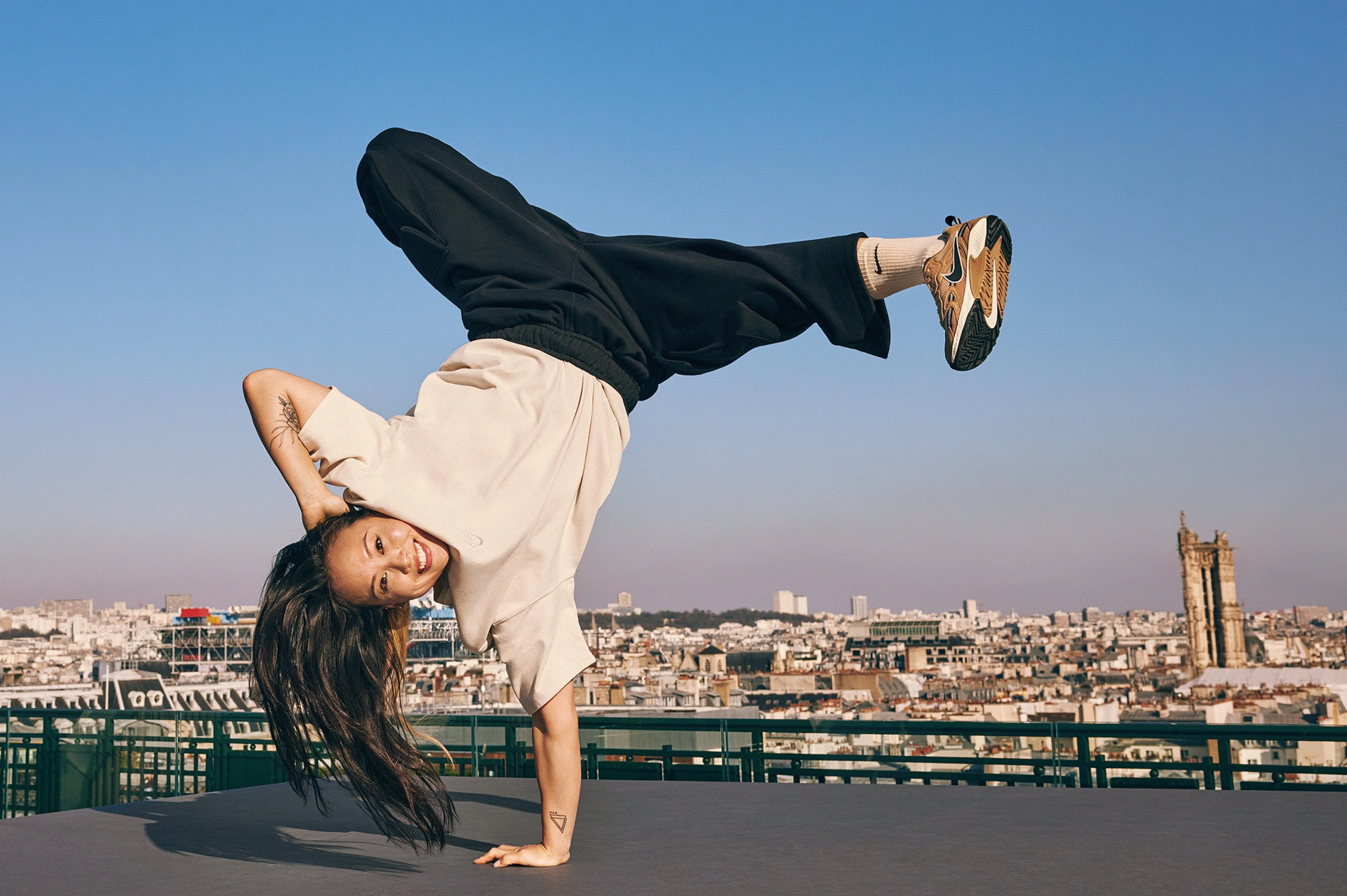 Sunny Choi does a one-handed handstand breakdancing move on a rooftop in Paris, France. Sunny wears black pants and a cream colored top from the Nike Sportswear collection, plus white Nike socks. The Paris city-scape with a bright blue sky is in the background.