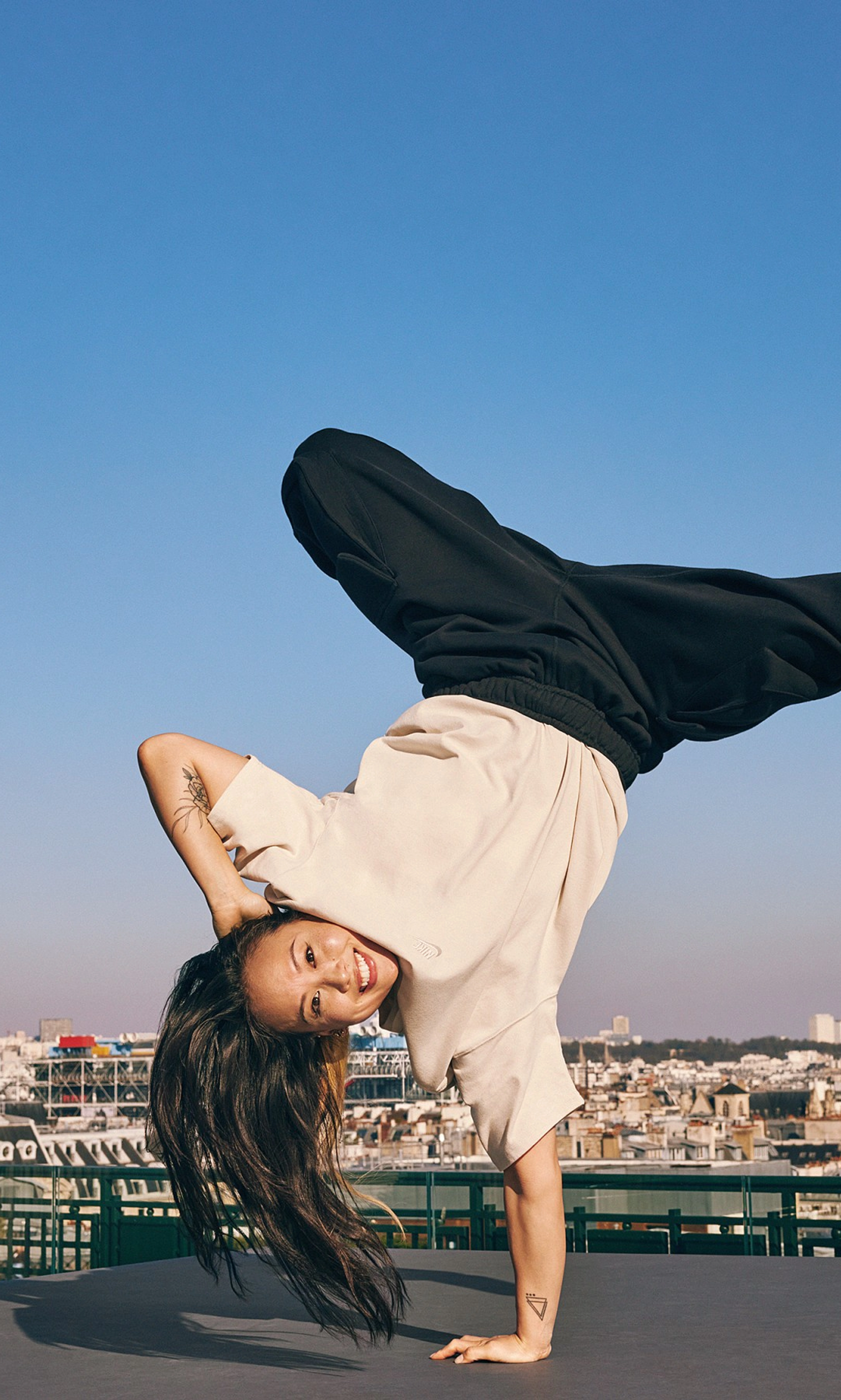 Sunny Choi does a one-handed handstand breakdancing move on a rooftop in Paris, France. Sunny wears black pants and a cream colored top from the Nike Sportswear collection, plus white Nike socks. The Paris city-scape with a bright blue sky is in the background.