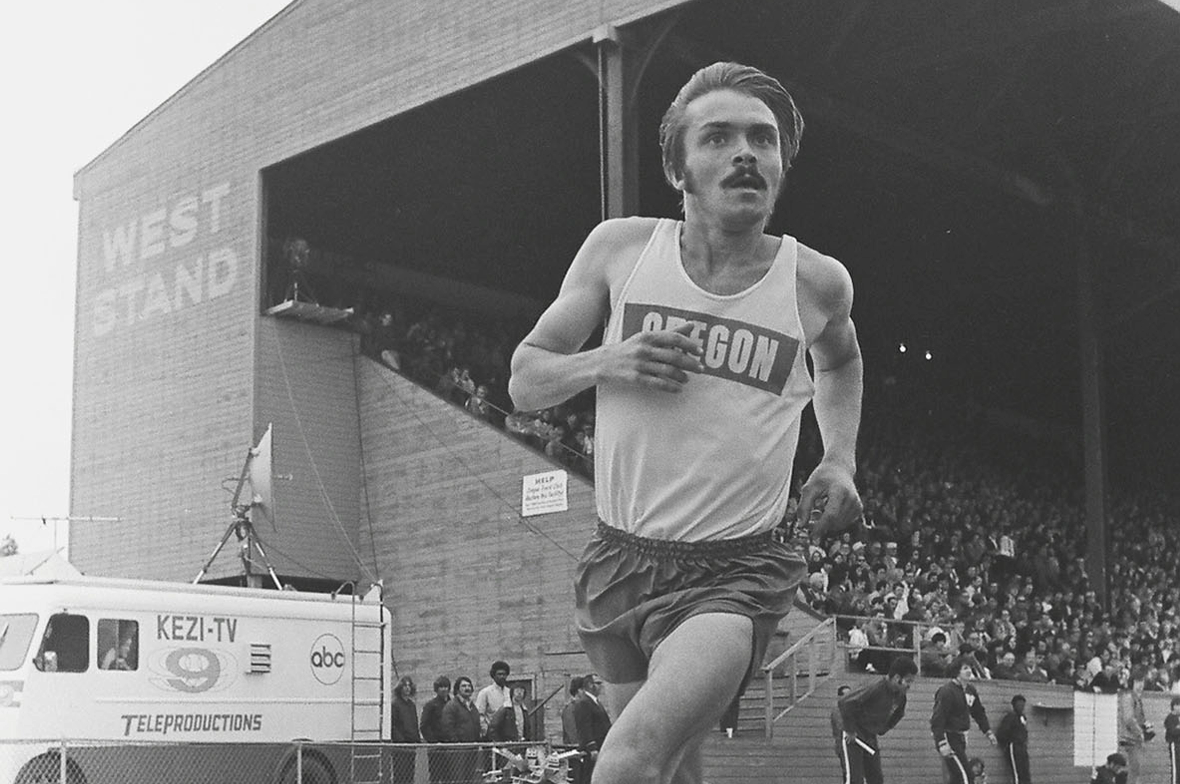 Black and white photo of Steve Prefontaine running on the track at Hayward Field at University of Oregon, Eugene, OR. Pre wears an Oregon running singlet with dark running shorts and looks down the track as he pumps his arms during a race.