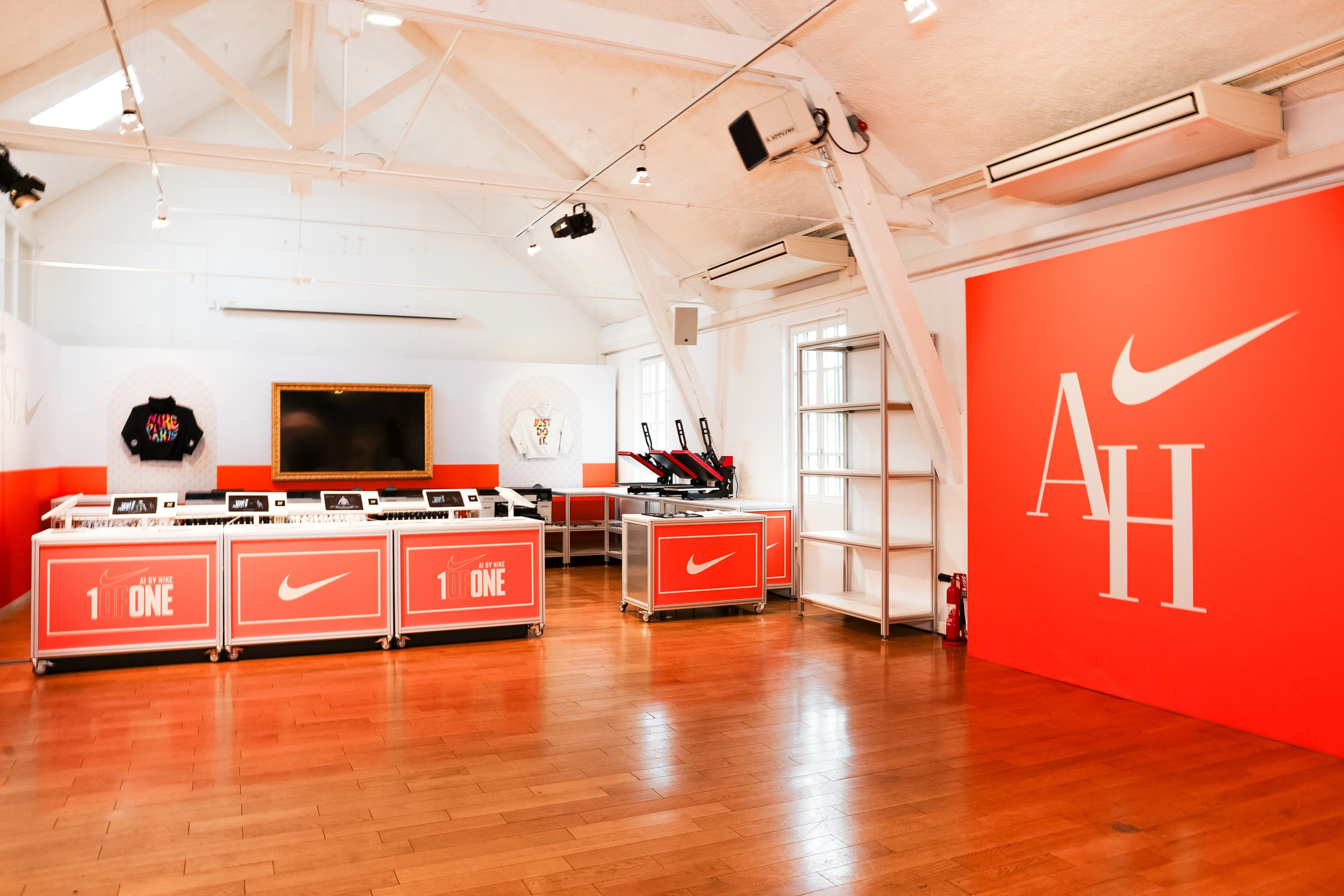 View into the AI customization space at the Nike Athlete House in Paris, France. To the right, there is a large orange wall with the AH logo. The orange customization stations sit on top of a polished wood floor, and we see customization screens and heat press units ready to be used.