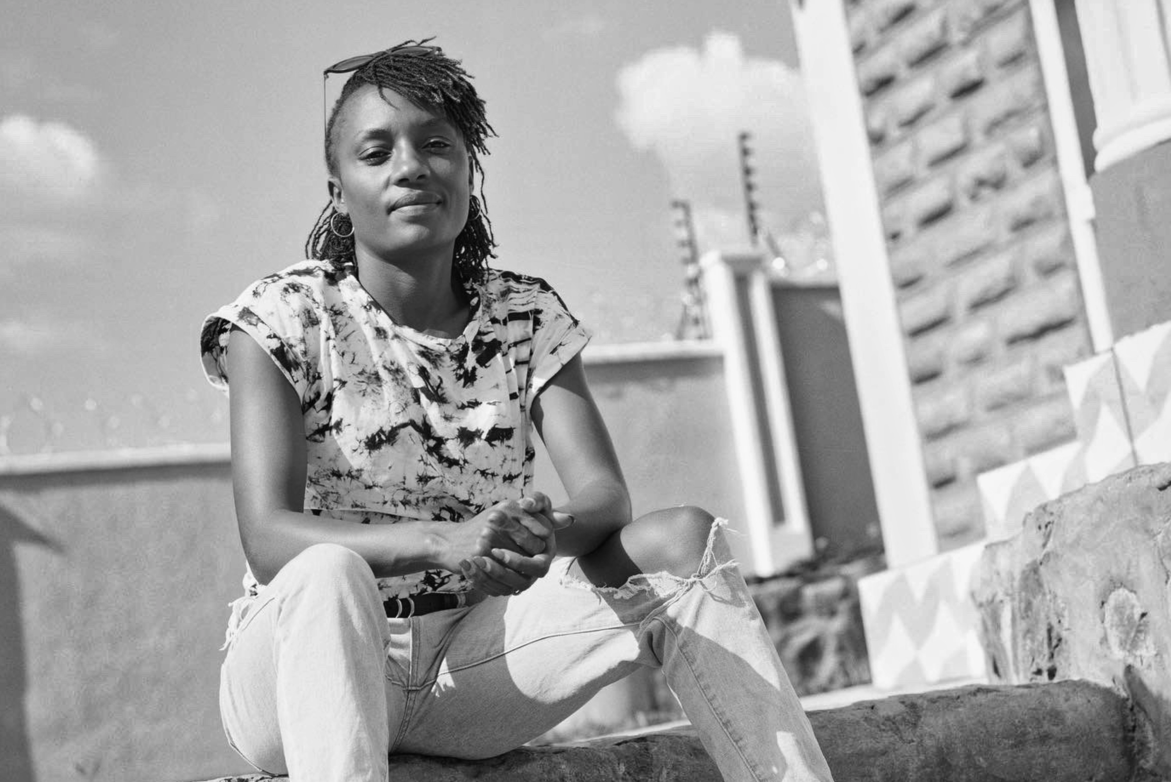 Black and white photo of Mary Ngugi in streetwear sitting on a concrete staircase in the city, her hands folded together with her elbows resting on her knees.