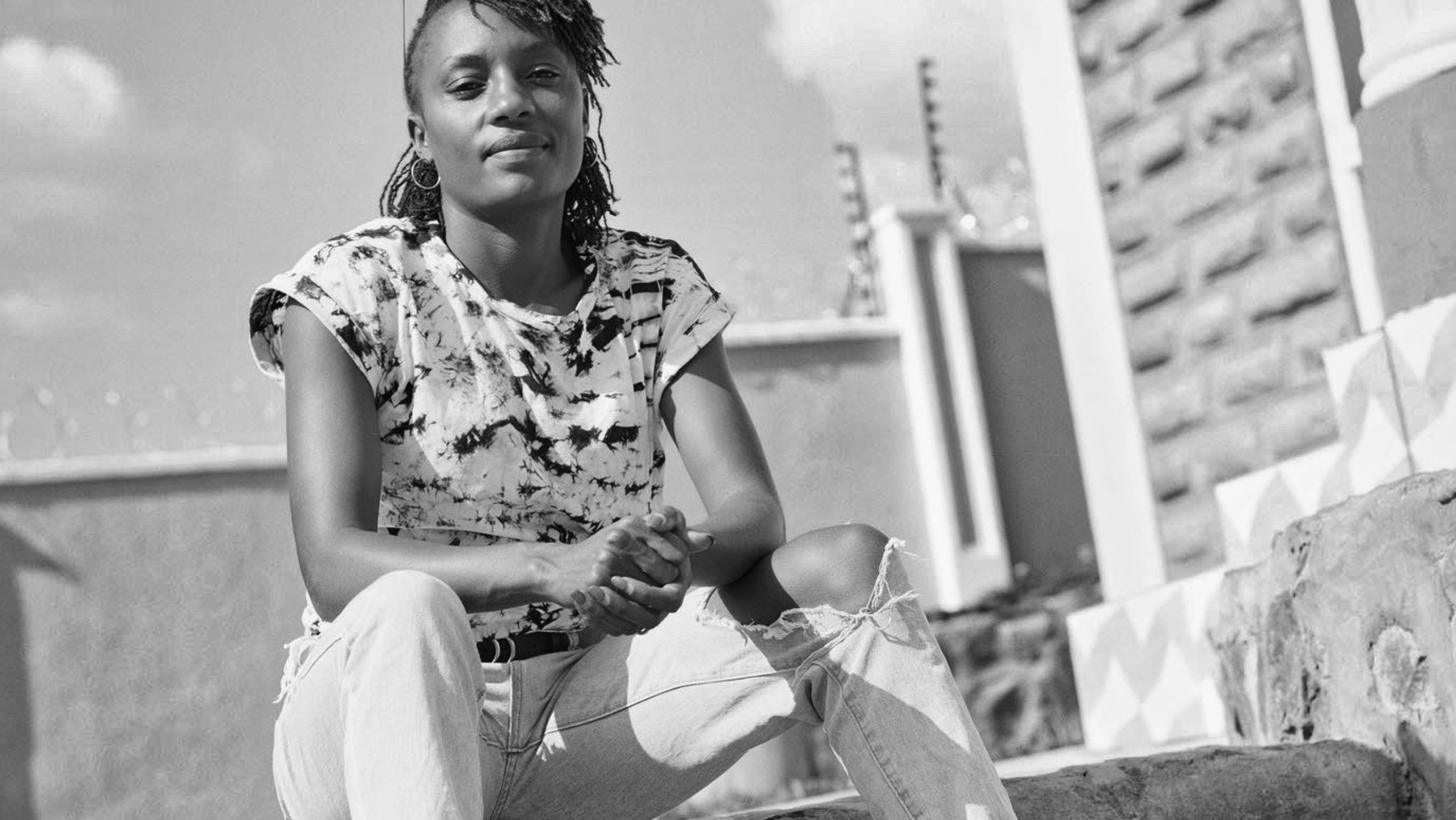 Black and white photo of Mary Ngugi in streetwear sitting on a concrete staircase in the city, her hands folded together with her elbows resting on her knees.