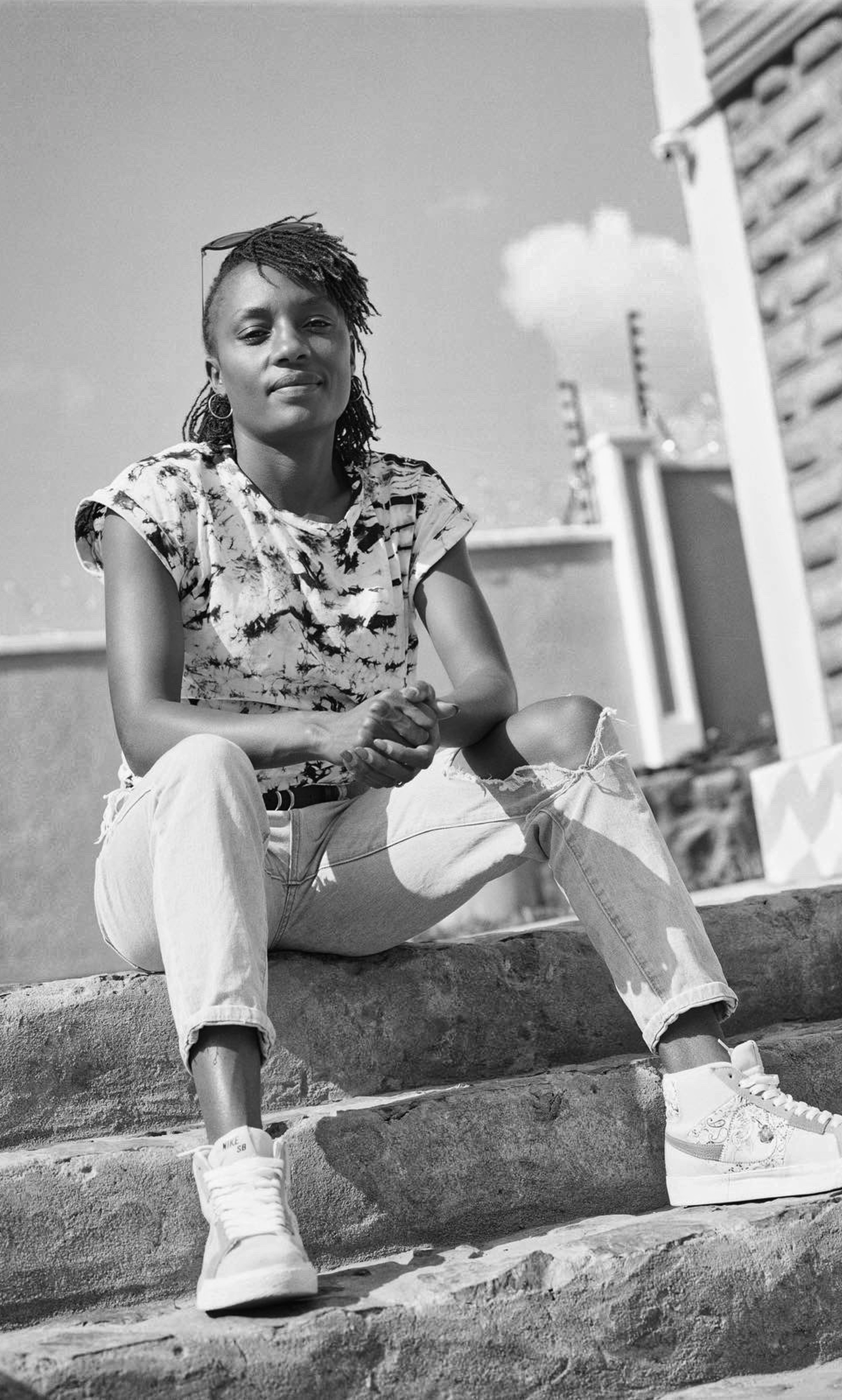 Black and white photo of Mary Ngugi in streetwear sitting on a concrete staircase in the city, her hands folded together with her elbows resting on her knees.