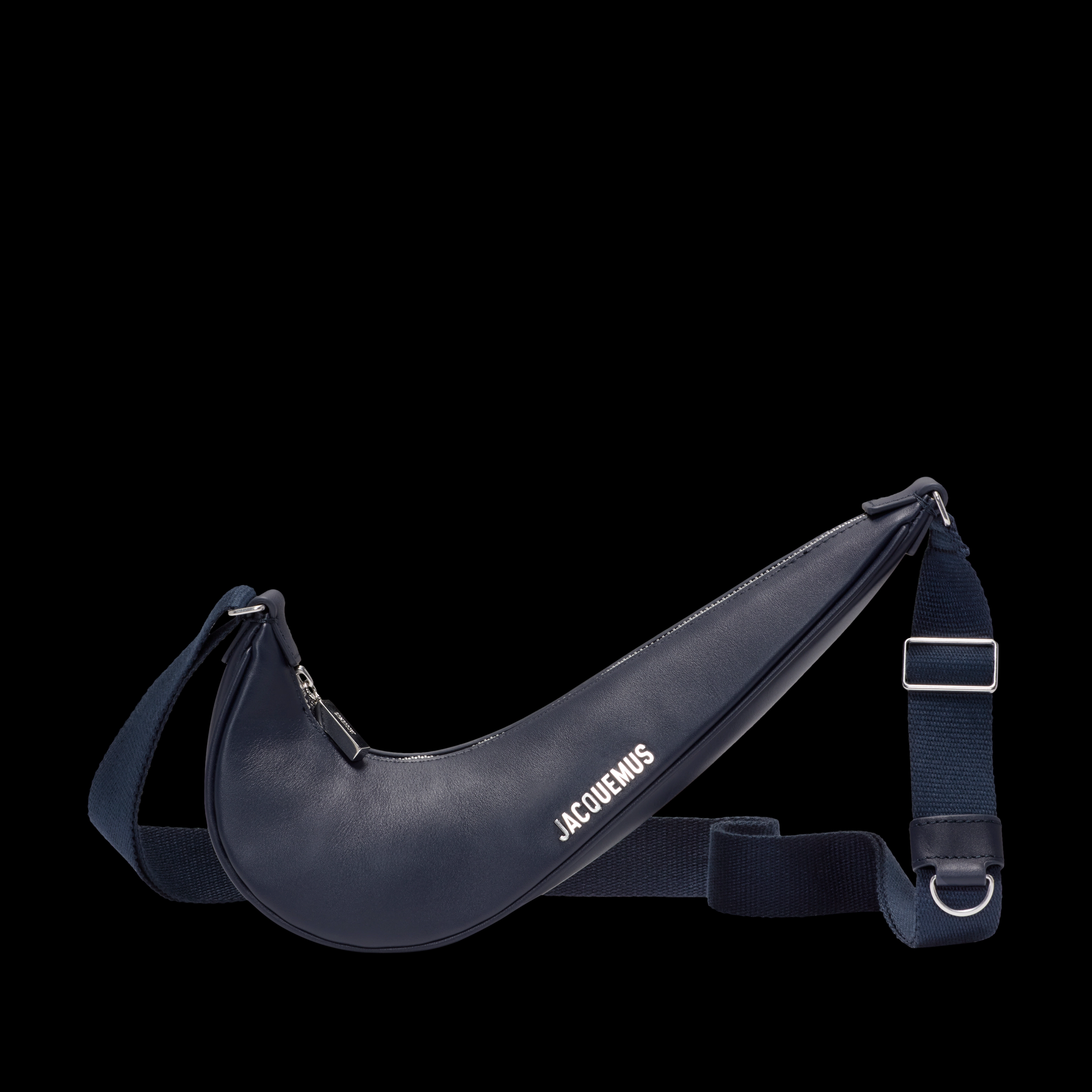 Front view of the Nike x Jacquemus Le Swoosh Crossbody Bag in midnight navy, with bright silver bag hardware and Jacquemus brand mark. The bag itself is in the shape of a Nike Swoosh, and includes a navy blue strap that is draped behind the body of the bag. The bag floats on a black background.