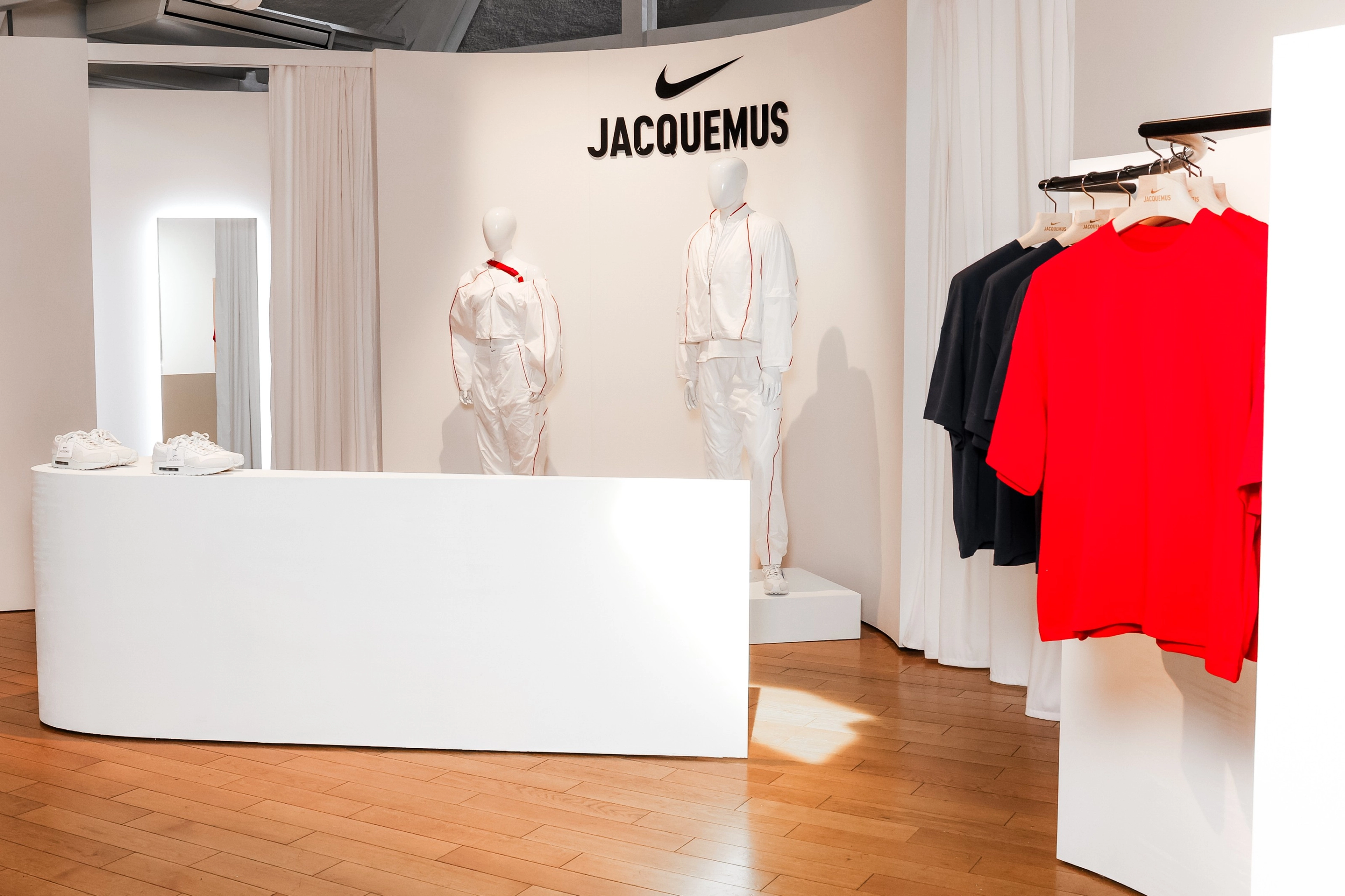 View into the Jacquemus Station at the Nike Athlete House in Paris, France.  We see the Nike X Jaquemus men's and women's track suits on white mannequins. Nike x Jacquemus short sleeve tees in Univserity red and black hang on a bar to the right. A curved white counter holds the Nike x Jacquemus Air Max 1 Sneaker in white. Fitting rooms can be seen in the background.