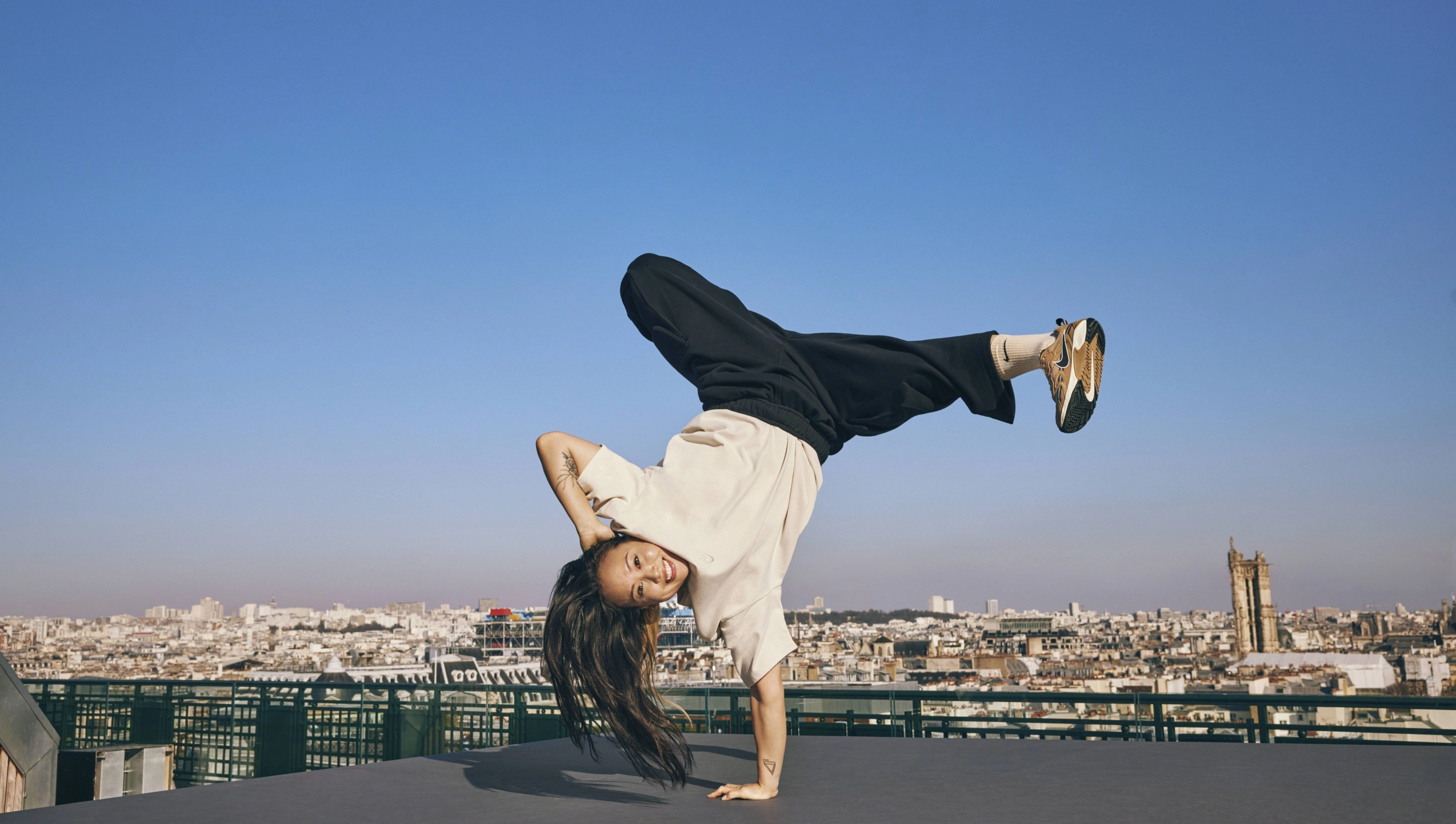 Sunny Choi does a one-handed handstand breakdancing move on a rooftop in Paris, France. Sunny wears black pants and a cream colored top from the Nike Sportswear collection, plus white Nike socks. The Paris city-scape with a bright blue sky is in the background.