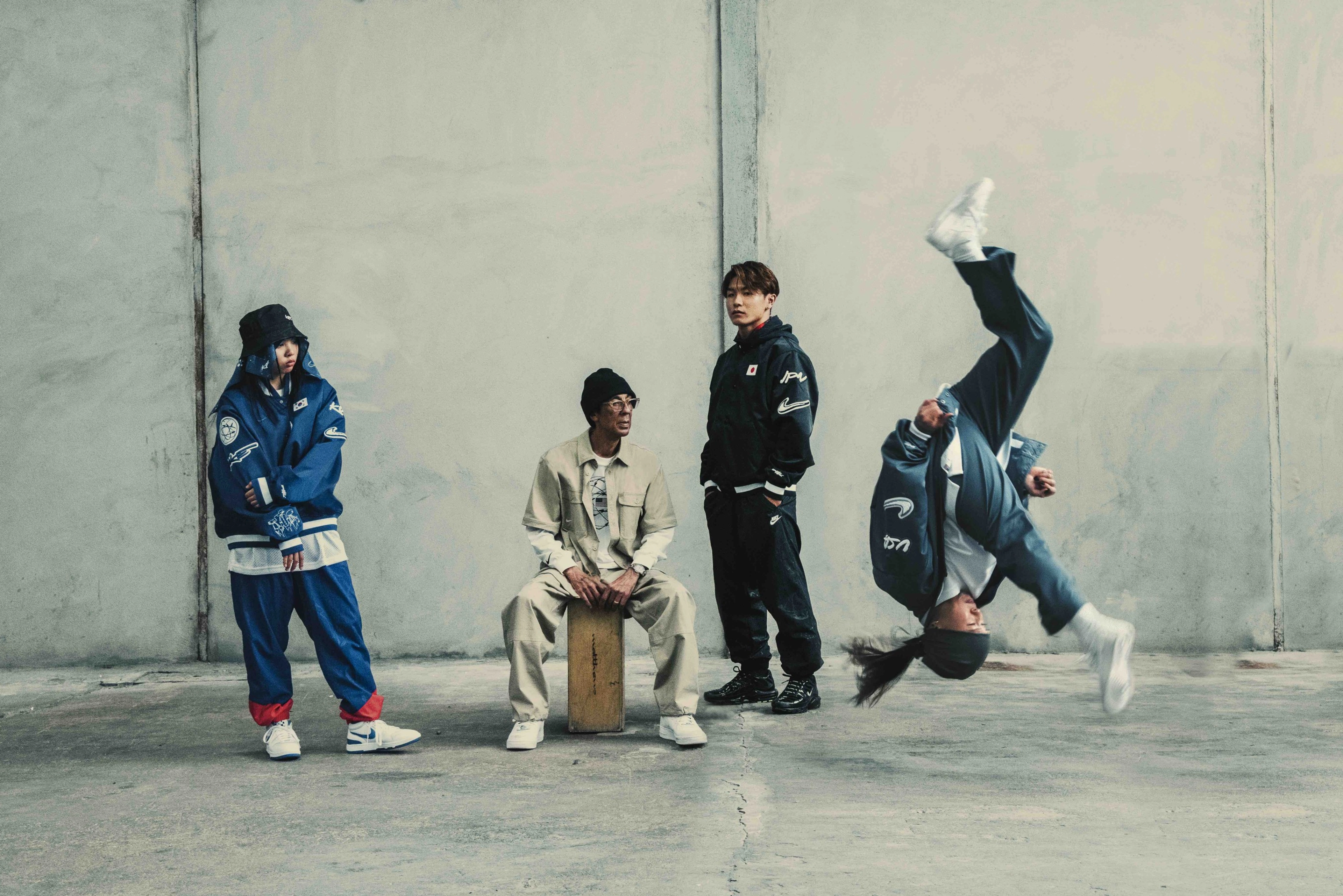 From left to right, JiYun Shun, Futura, and Shigeyuki Nakari watch as Sunny Choi does a front flip during a breakdance in white Nike Jam shoes. The breakdancers are located in a concrete space with grey floors and walls, and all wear attire from the Nike Breaking Collection.