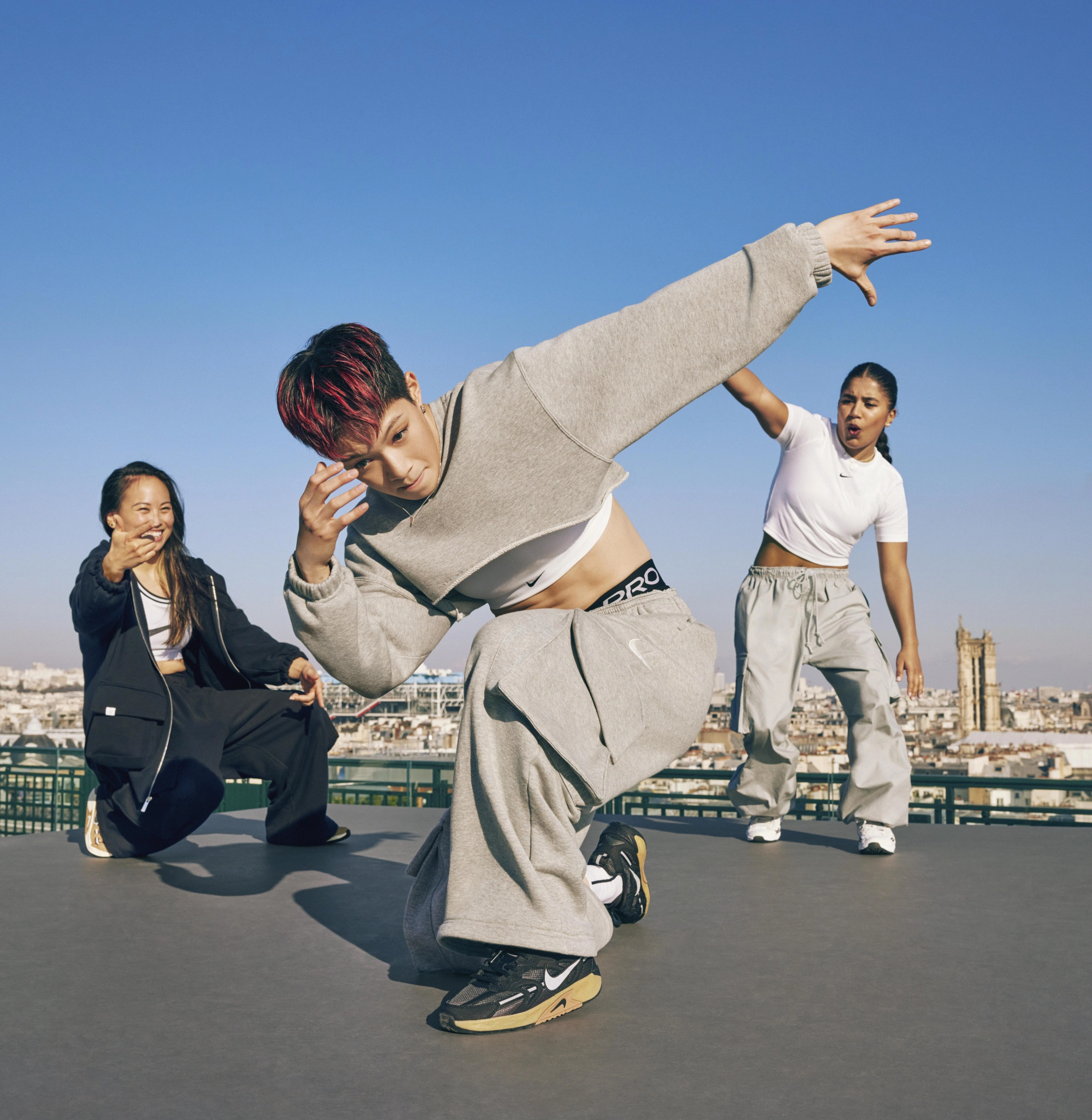 From left to right, Sunny Choi, Yell-Yeri Kim, and India Sardjoe appear on a rooftop wearing Nike Jam sneakers and a variety of Nike Sportswear clothing. The Paris cityscape is in the background, with a glimpse of the Centre Pompidou.