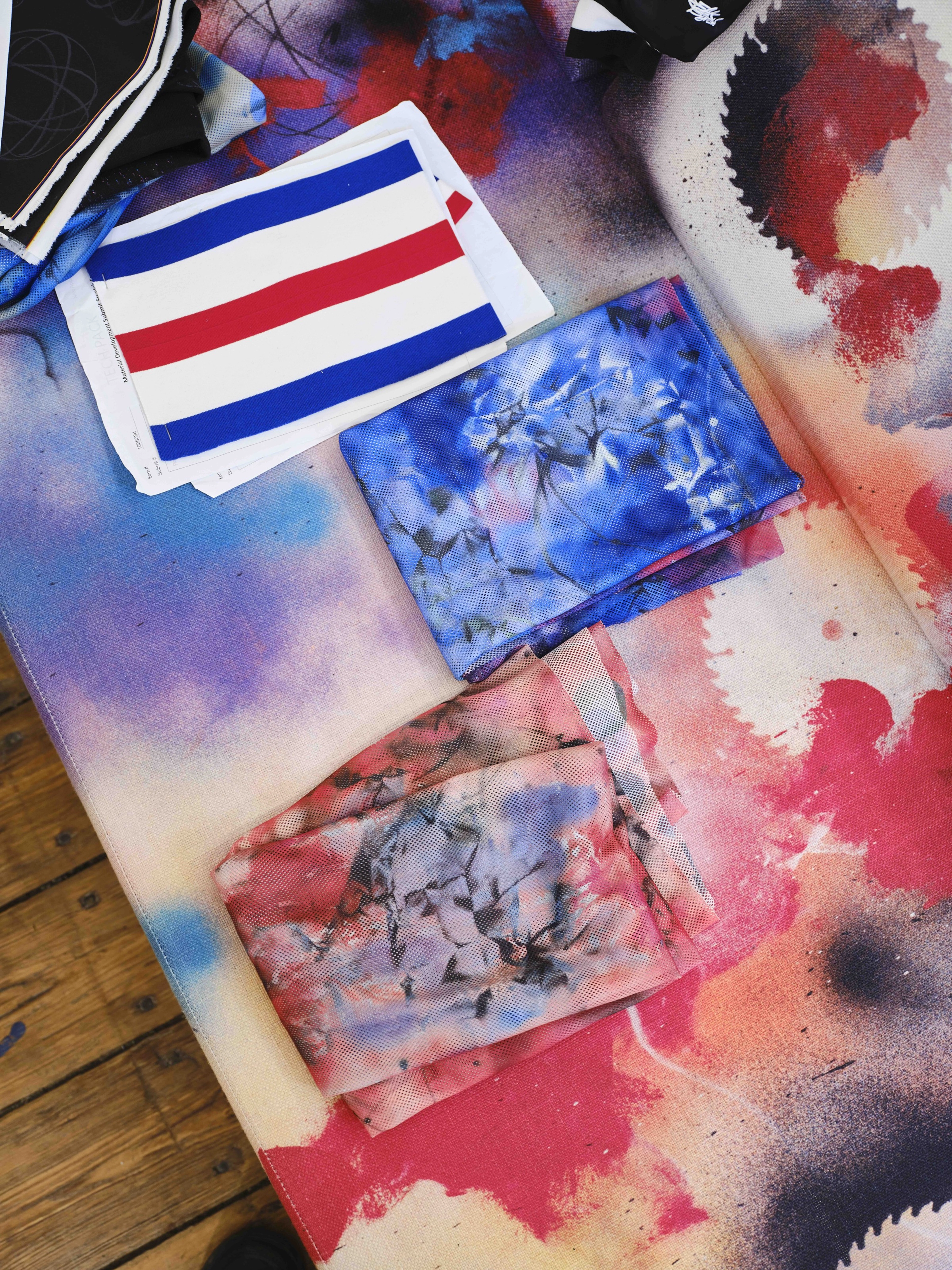 Detail view of painted cloth material and swatches, featuring bright reds, pinks, blues and purples. The material sits on a wood floor.