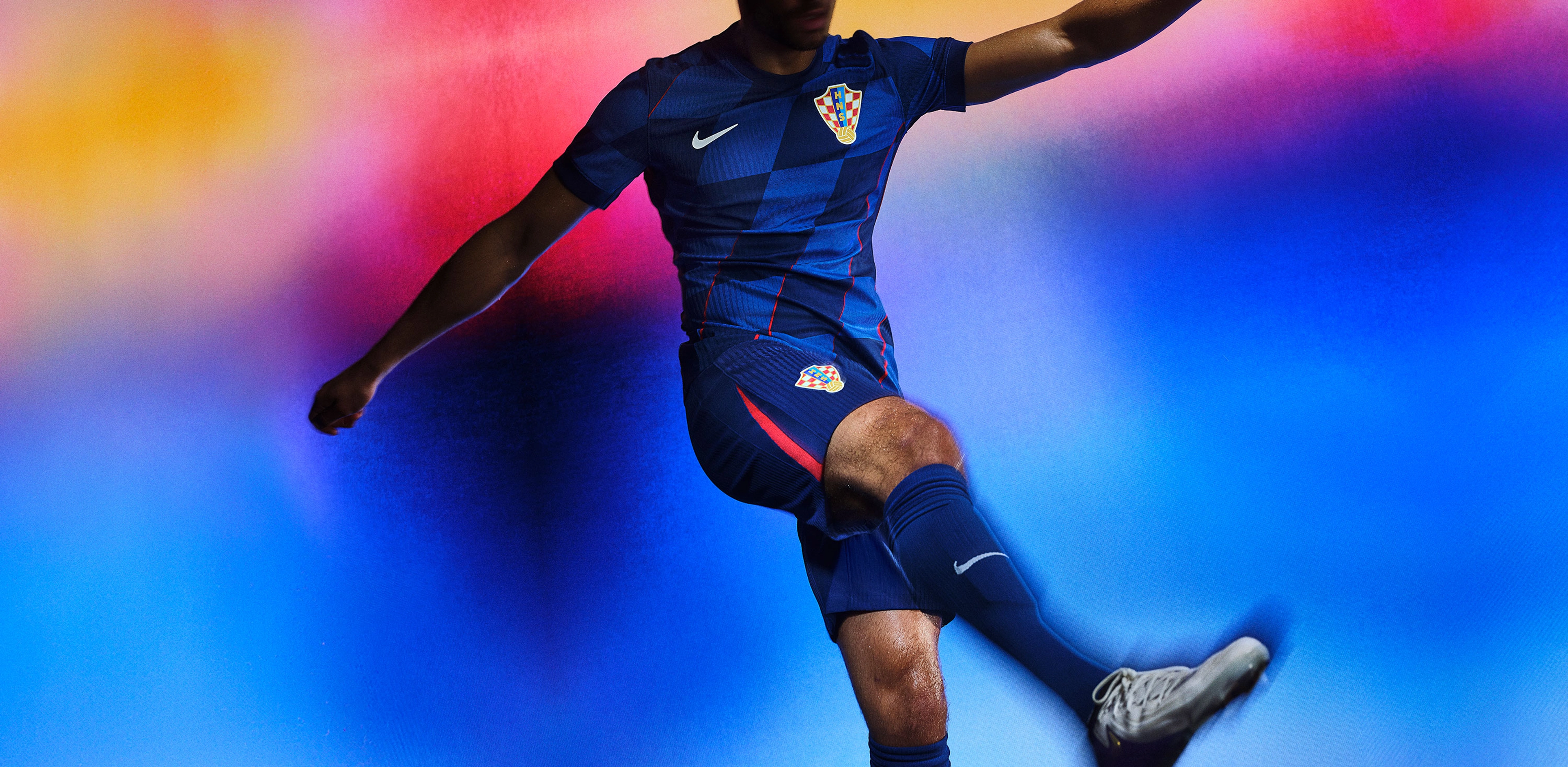 Athlete in a kicking pose wearing a soccer jersey with a blue rhombus pattern with bright red lines intersecting them, as well as matching blue shorts with a bright red stripe on the side, each with a Nike swoosh and team logo on the chest and legs. They are also wearing matching blue socks.