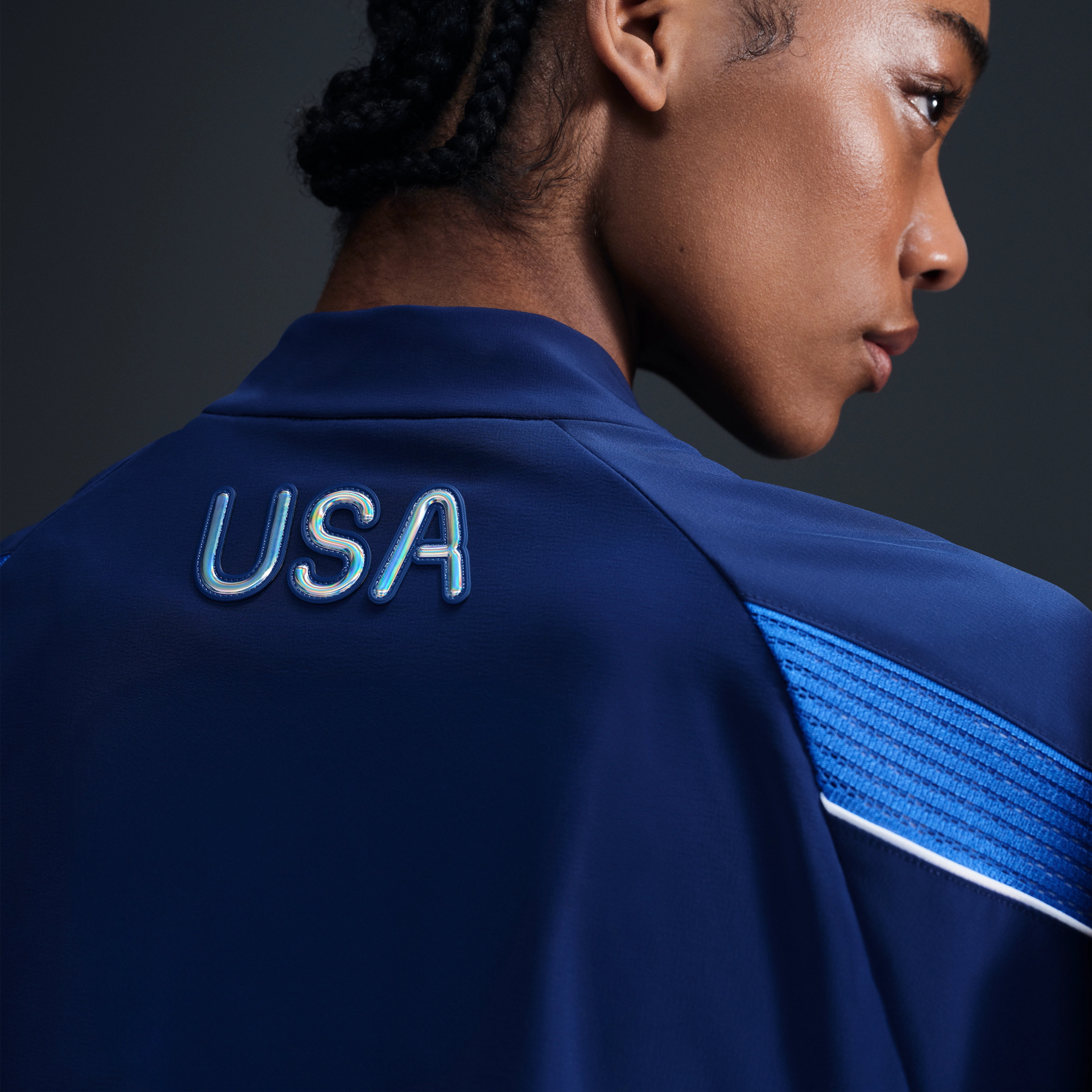 Back view of athlete wearing Women's Medal Ceremony Jacket in the exclusive bright blue Team USA colorway.  USA is emblazoned on the back of the jacket in iridescent silver letters. She stands against a dark background.