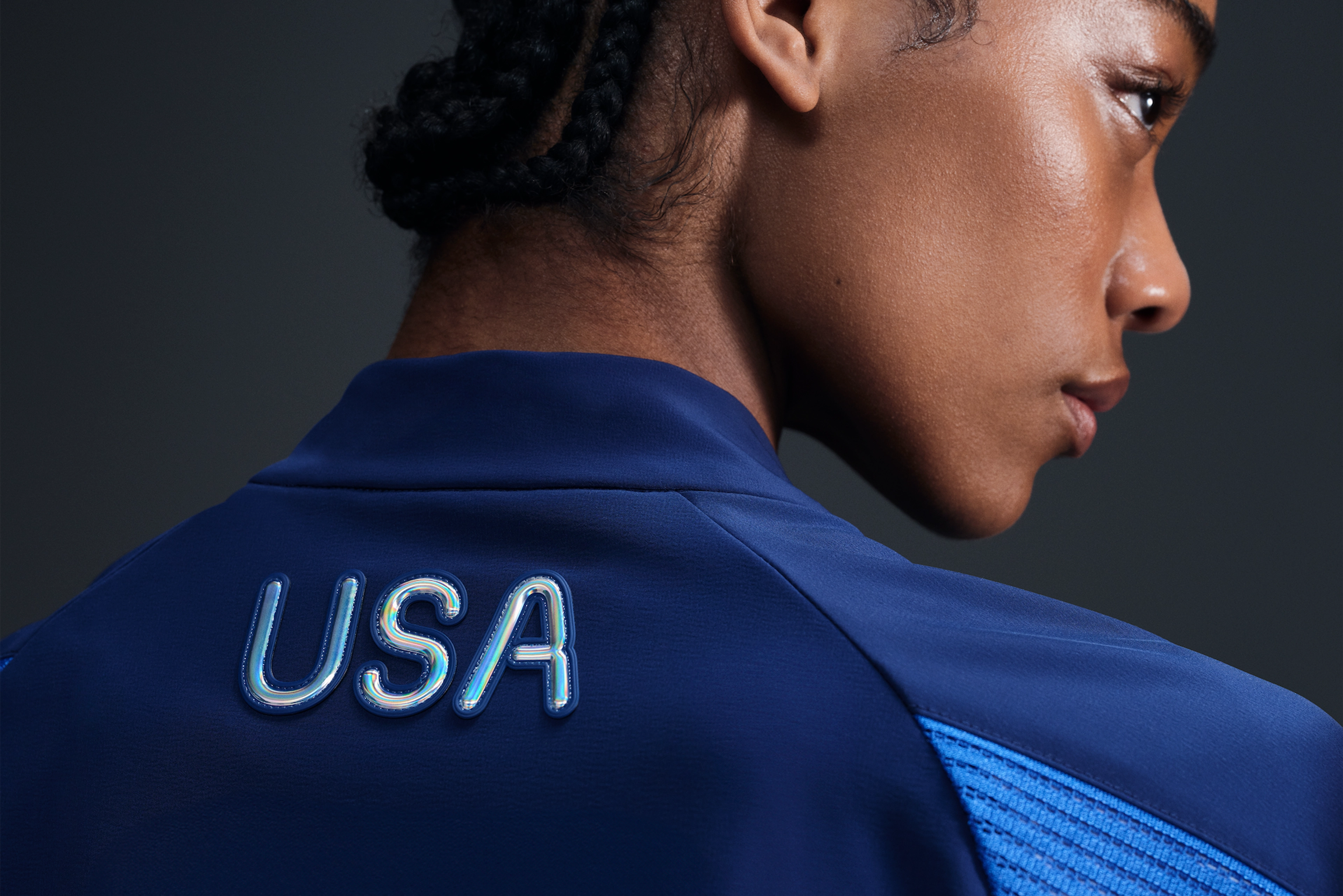 Back view of athlete wearing Women's Medal Ceremony Jacket in the exclusive bright blue Team USA colorway. USA is emblazoned on the back of the jacket in iridescent silver letters. She stands against a dark background.