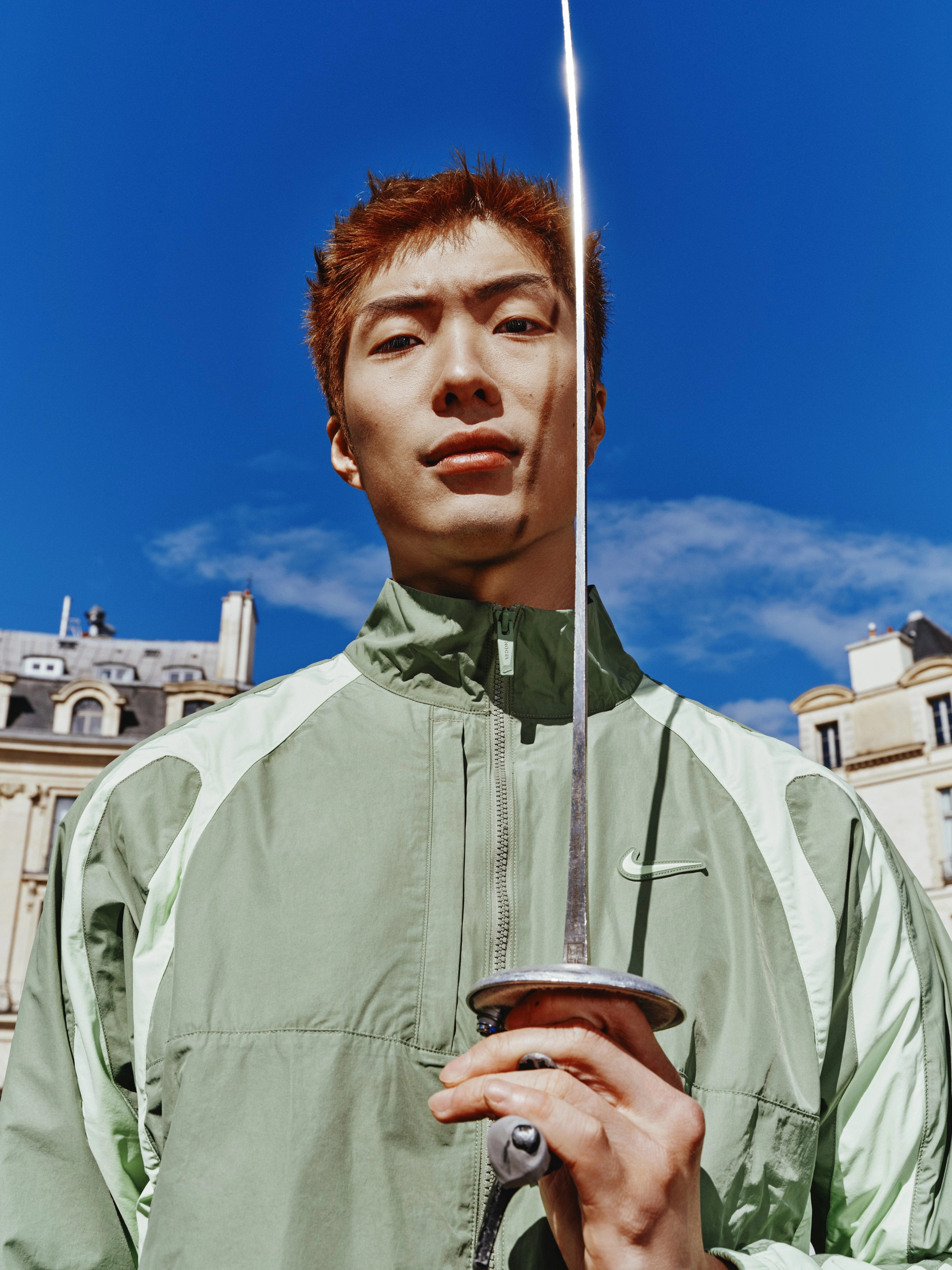 Cheung Ka Long holds his fencing foil upwards in front of his face, while looking directly at the camera. We see only the top half of his body, and he is wearing the NOCTA Northstar Nylon Track Jacket in Oil Green. There is a bright blue sky behind him, and glimpses of a couple of buildings.