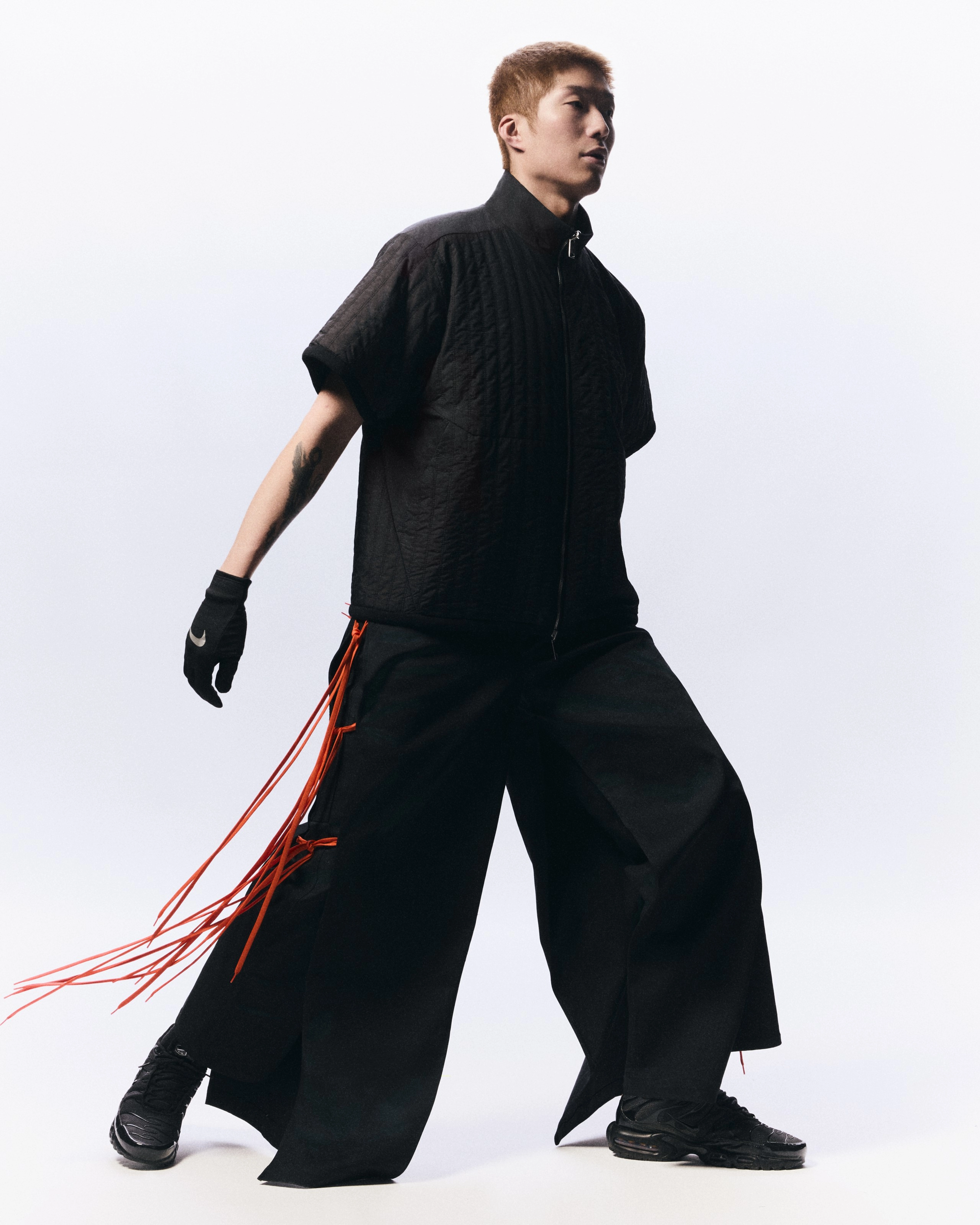 Full body portrait of Cheung Ka Long as he strides across a white background. He wears oversized black pants with red ties, a black short-sleeve zip up shirt, black Nike footwear, and black gloves with a reflective Swoosh on the back of the hand.