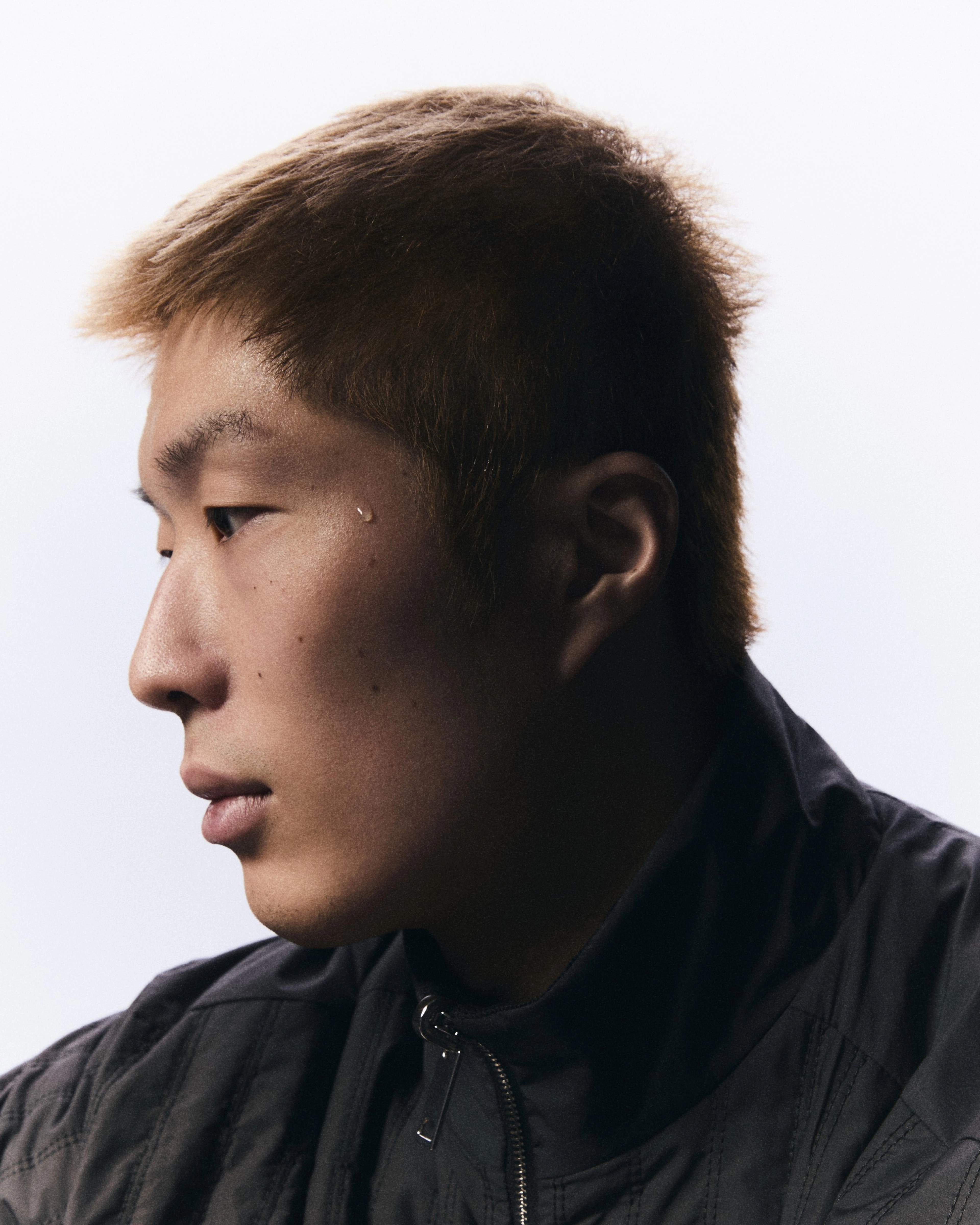 Side view portrait of Cheung Ka Long, facing to the left. His face is framed against a white background, and he wears a black jacket.  His face glows in the light, while his neck and ear is in shadow.
