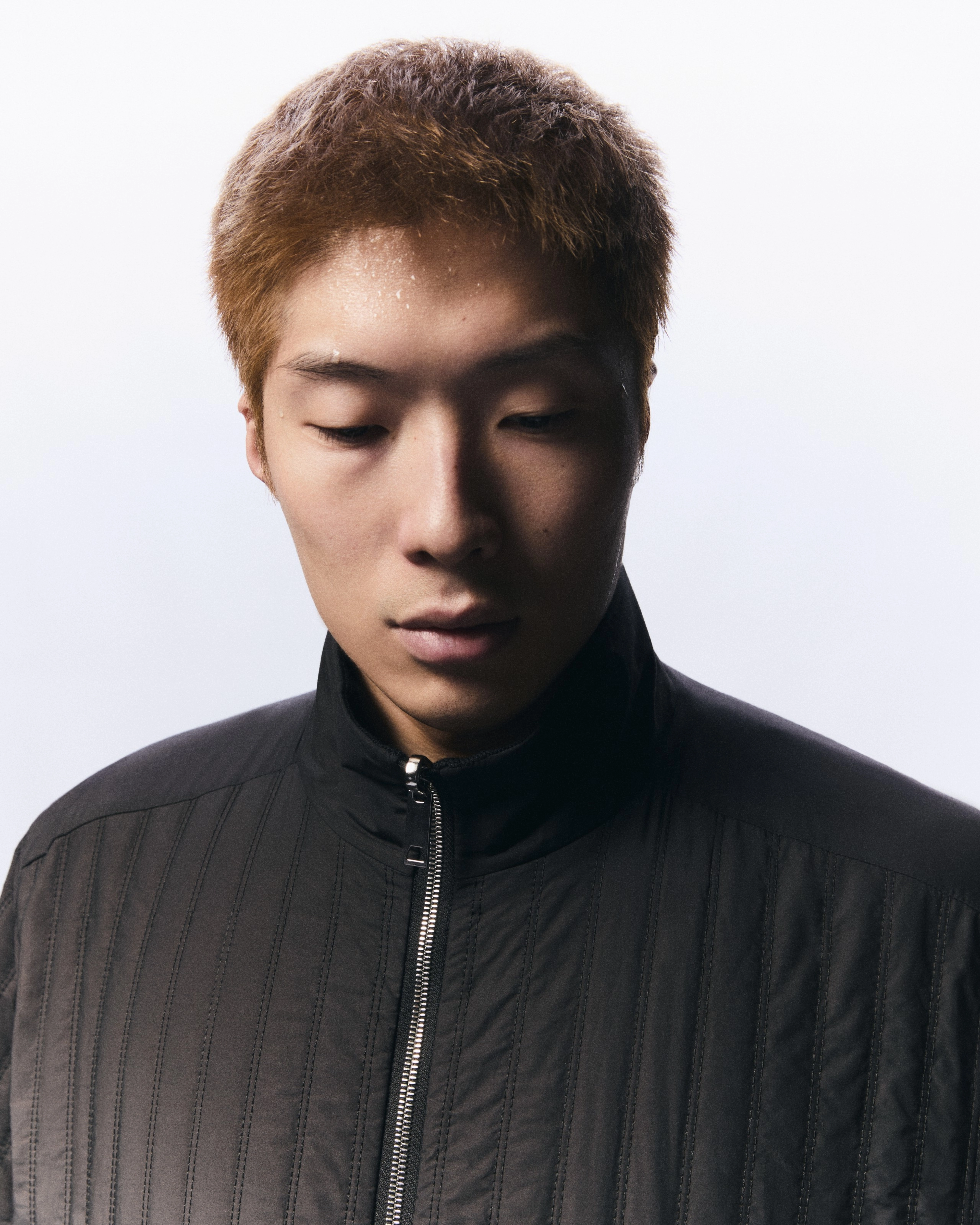 Upper body portrait of Cheung Ka Long. He faces the camera, but looks downwards, and lighting highlights the left side of his face. He wears a black zippered jacket and is framed by a white background.