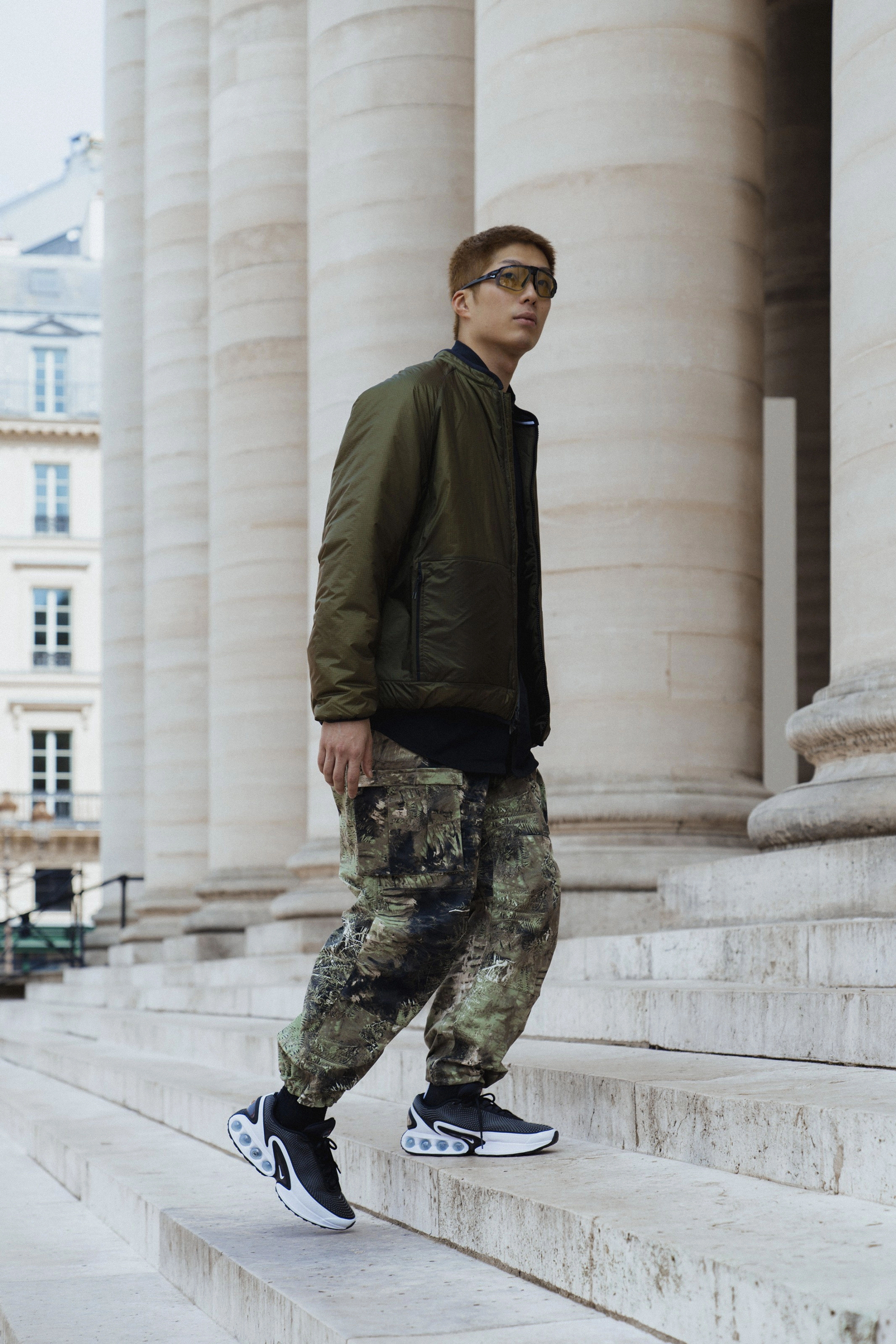 Cheung Ka Long walks up stone steps, in front of large stone pillars. He is wearing the Nike ACG "Smith Summit" all over print pants, with black Air Max DN footwear, and an olive green jacket. He wears Nike eyewear with wrapping lenses and a bold black frame. He looks off to the right.