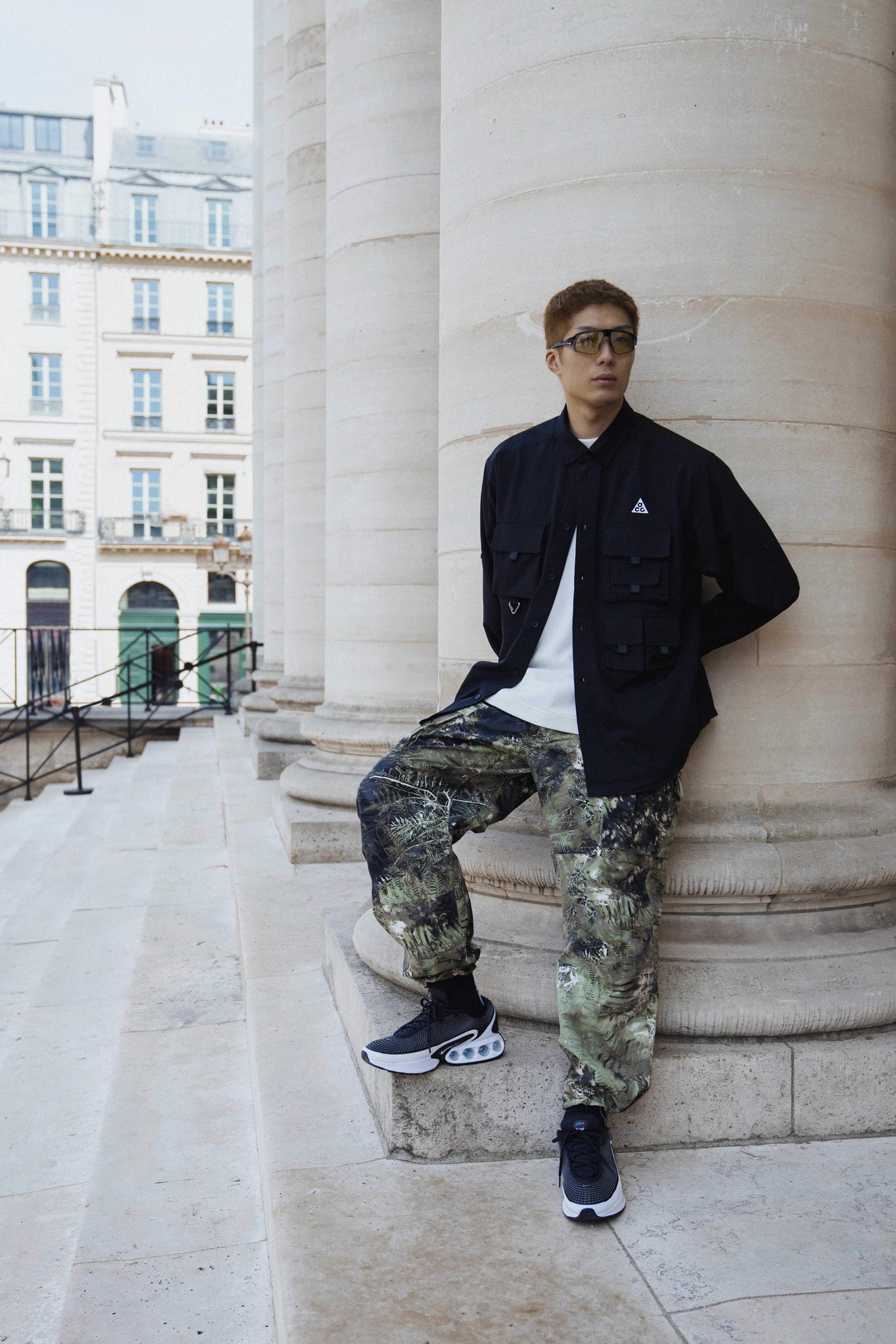 Full body view of Cheung Ka Long, leaning against the base of a large stone pillar. He wears the Nike ACG "Smith Summit" pant in an all over green camo print, black Air Max DN footwear, and a black Nike ACG Dri-FIT UV "Devastation Trail" shirt, with a white tee peeking out from underneath. Long wears Nike glasses with a wrapping lens and black frames. In the background we can see a glimpse of european buildings with green doors.