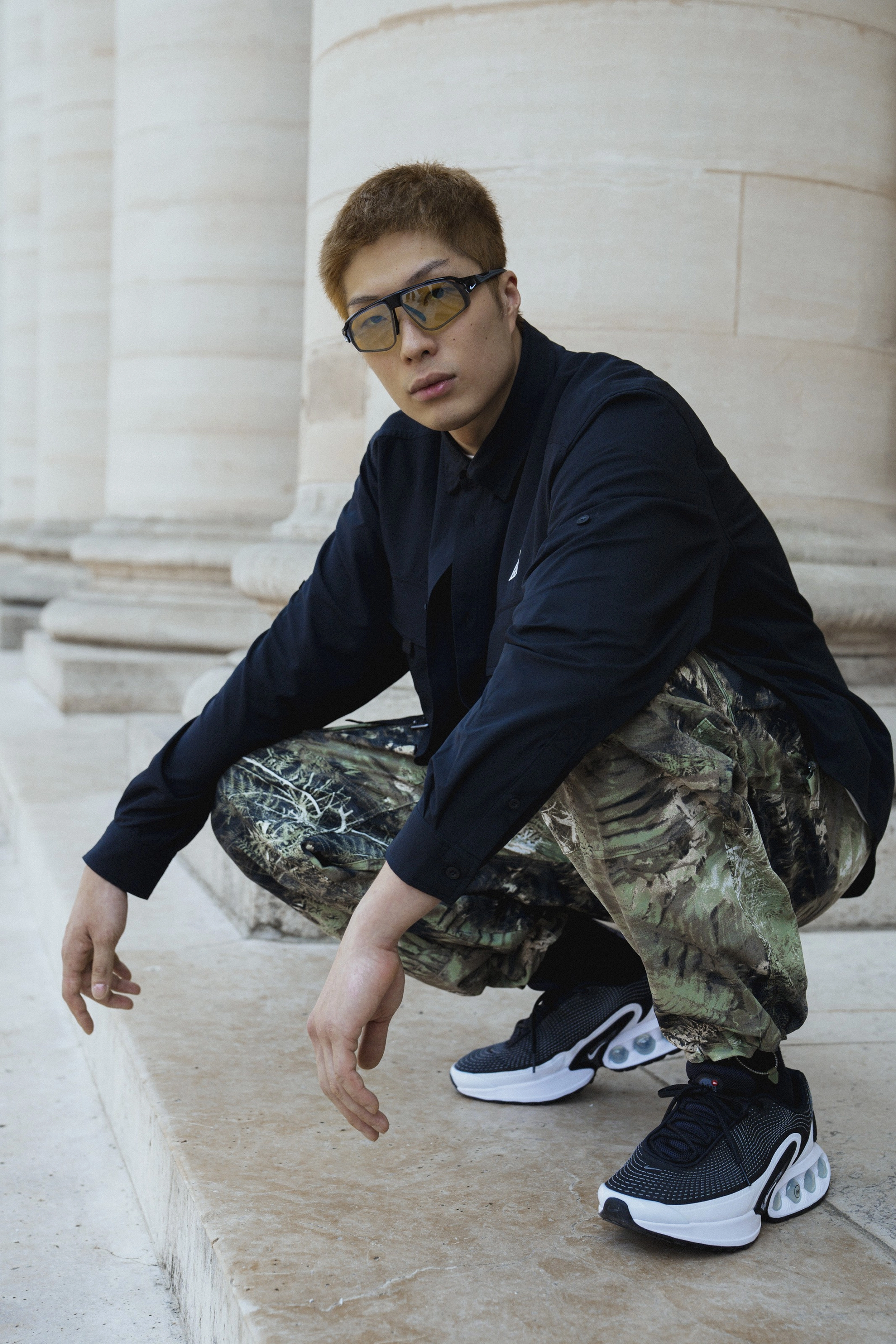 Full body portrait of Cheung Ka Long, crouching in front of large stone pillars.  He is wearing a pair of Air Max DN shoes, Nike ACG "Smith Summit" pants with a green all-over camo print, and a black Nike ACG Dri-FIT UV "Devastation Trail" long sleeve shirt. Long looks directly at the camera, wearing a pair of Nike glasses with wrapping lenses and bold black frames.