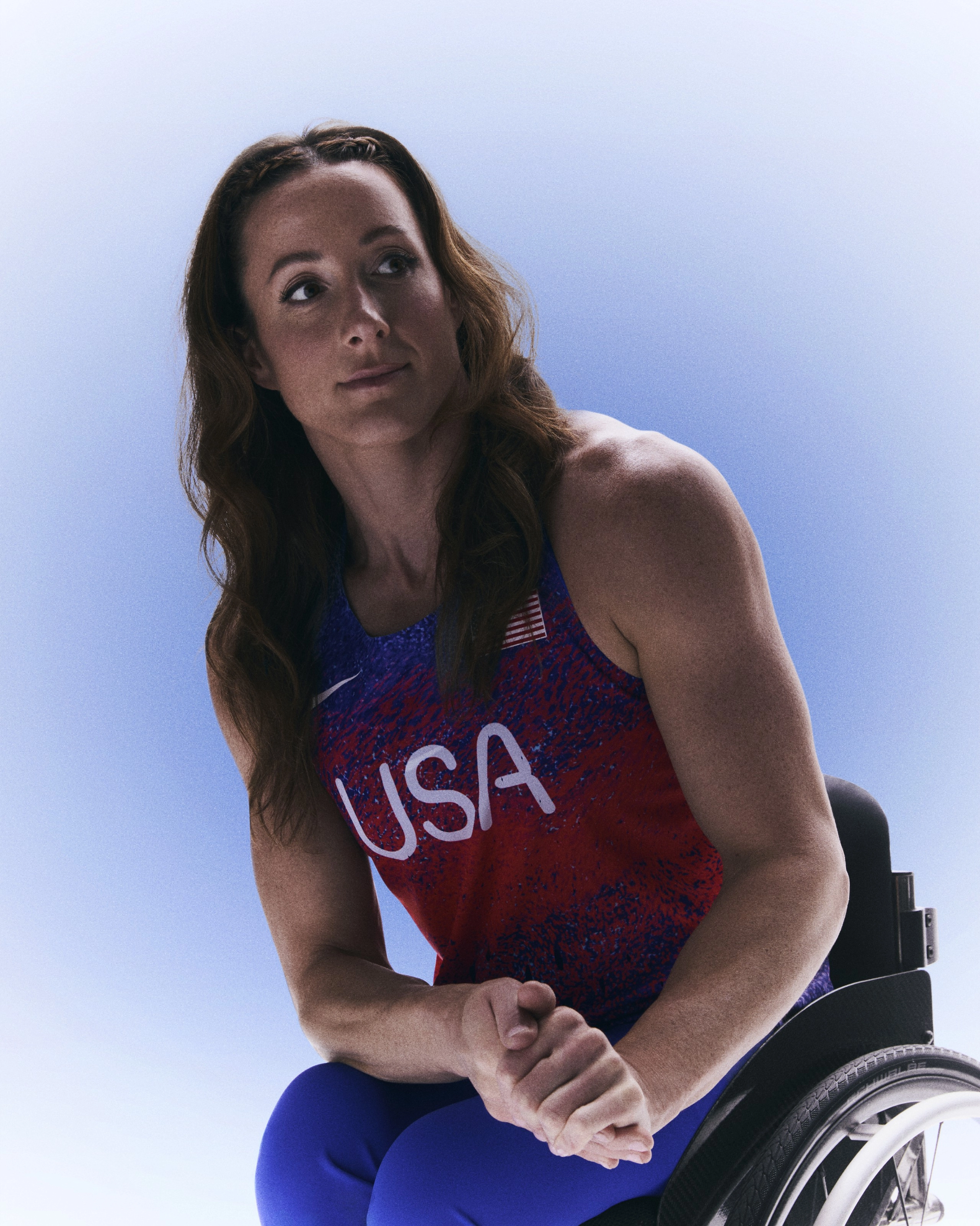 Portrait of Tatyana McFadden in her Team USA Running singlet with her hair down on her shoulders. Tatyana looks to the right while holding her hands together, sitting in her wheelchair against a blue gradient background.