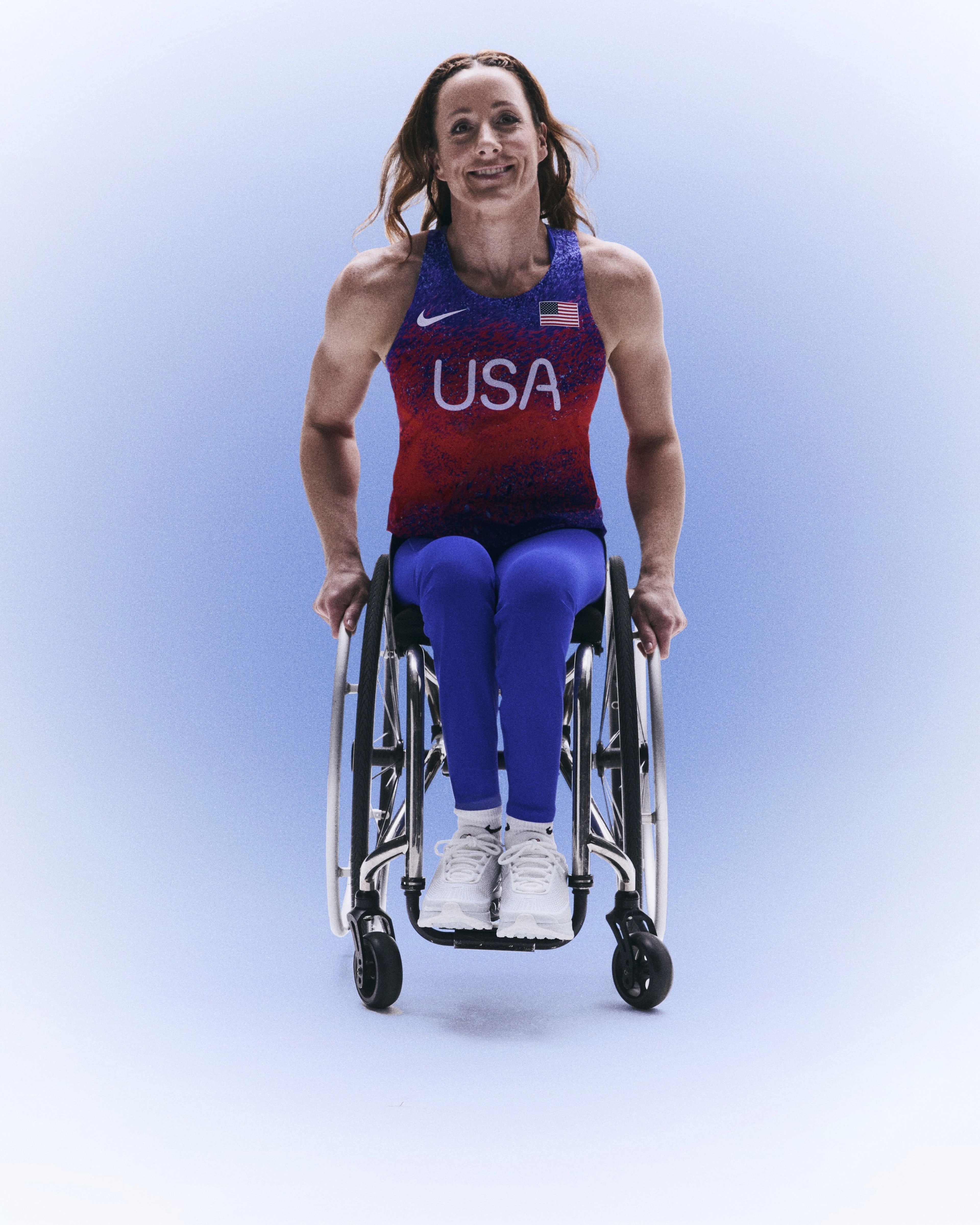 Full body portrait of Tatyana McFadden wearing her Team USA track and field uniform, with her hands resting on the hand rails of her wheelchair.  Tatyana smiles directly at the camera with her hair blowing slightly back, against a blue gradient background.
