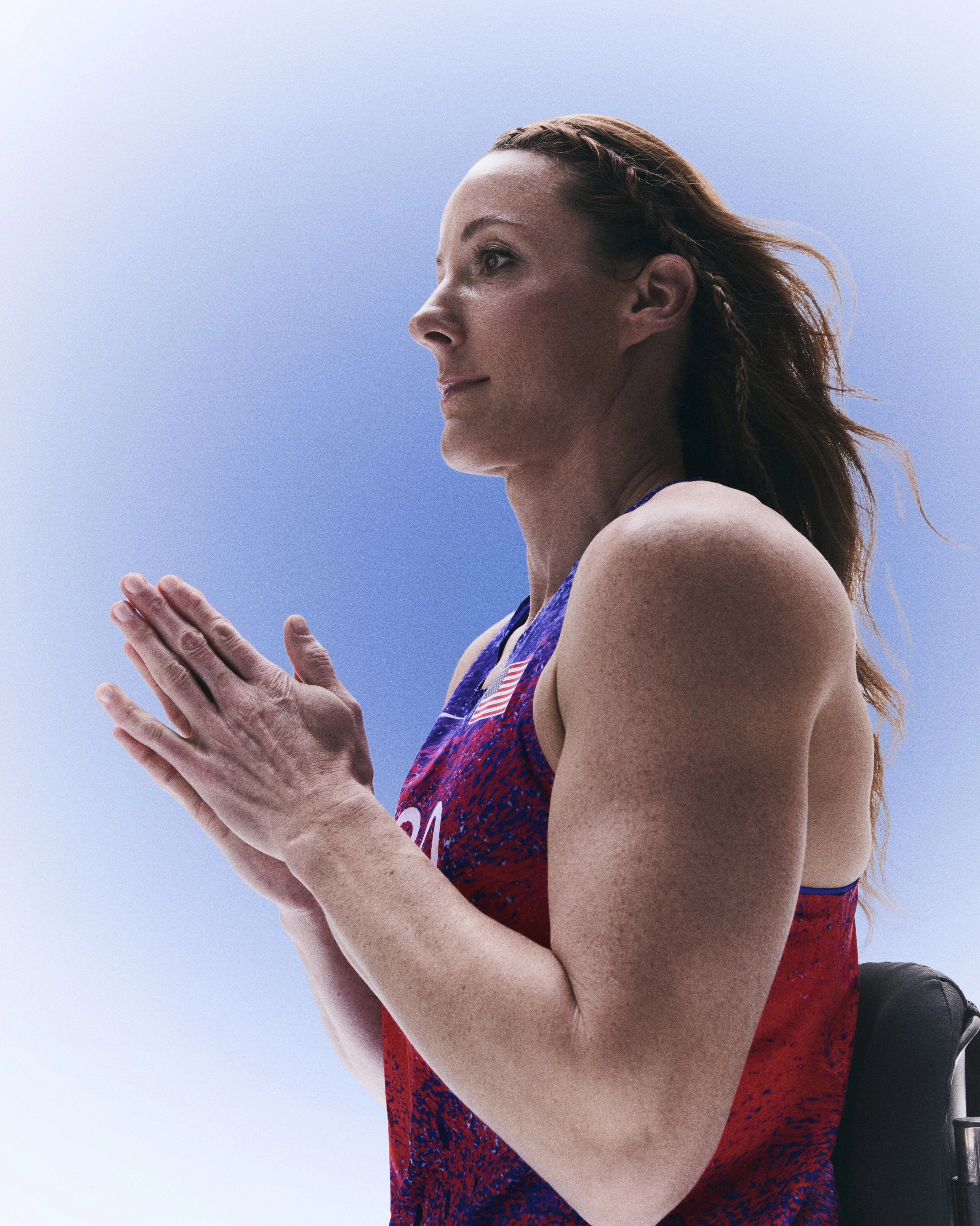 Side view of Tatyana McFadden pressing her hands together, while facing left. Tatyana wears the Team USA track and field singlet, and her hair is loose with a single braid across the front. She appears against a blue gradient background.