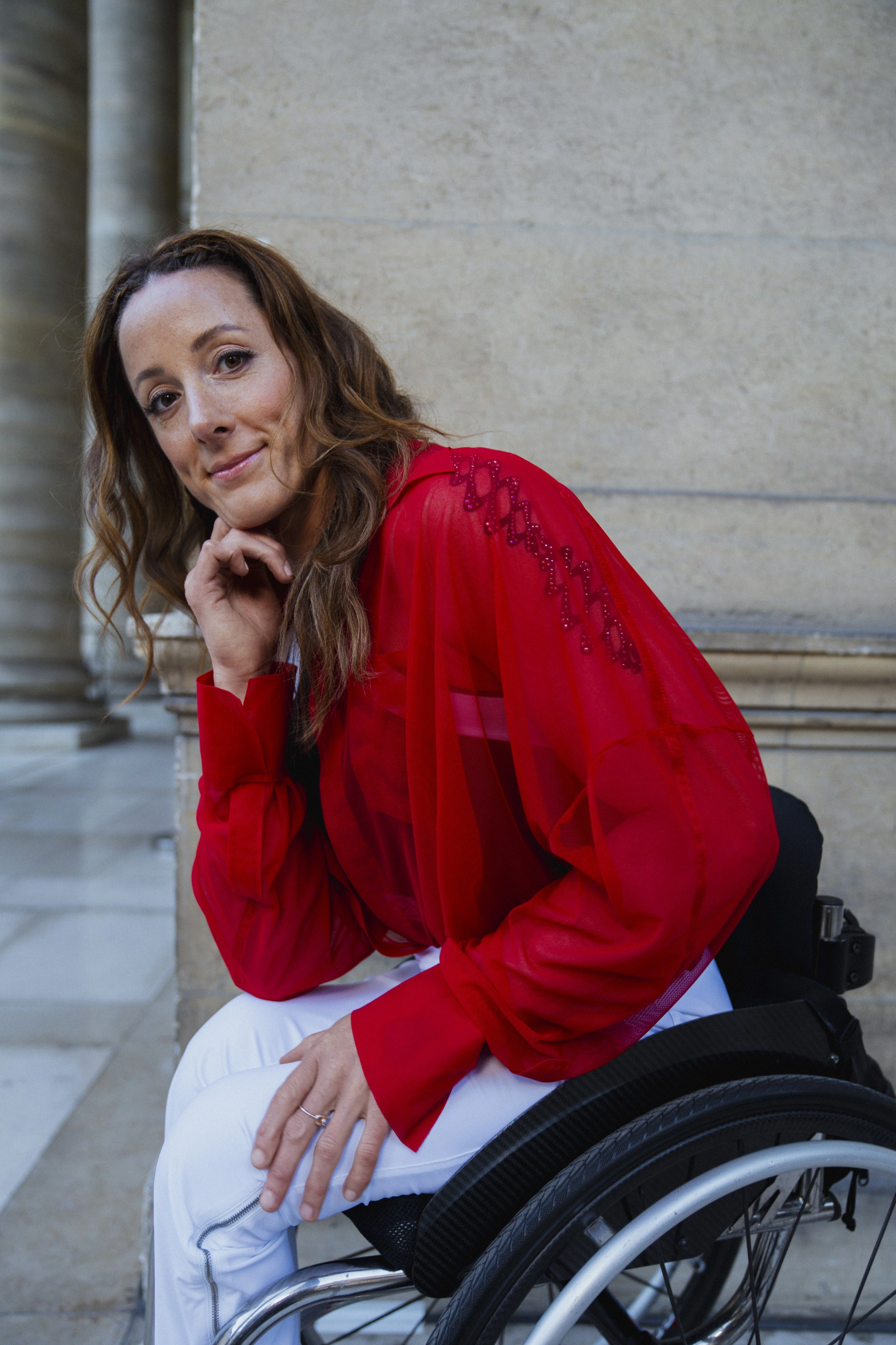 Tatyana McFadden leans forward, resting her chin on her hand, as she sits in her wheelchair among architectural pillars in a European setting. Tatyana wears a red blouse and white track pants, and her hair is down over her shoulders.
