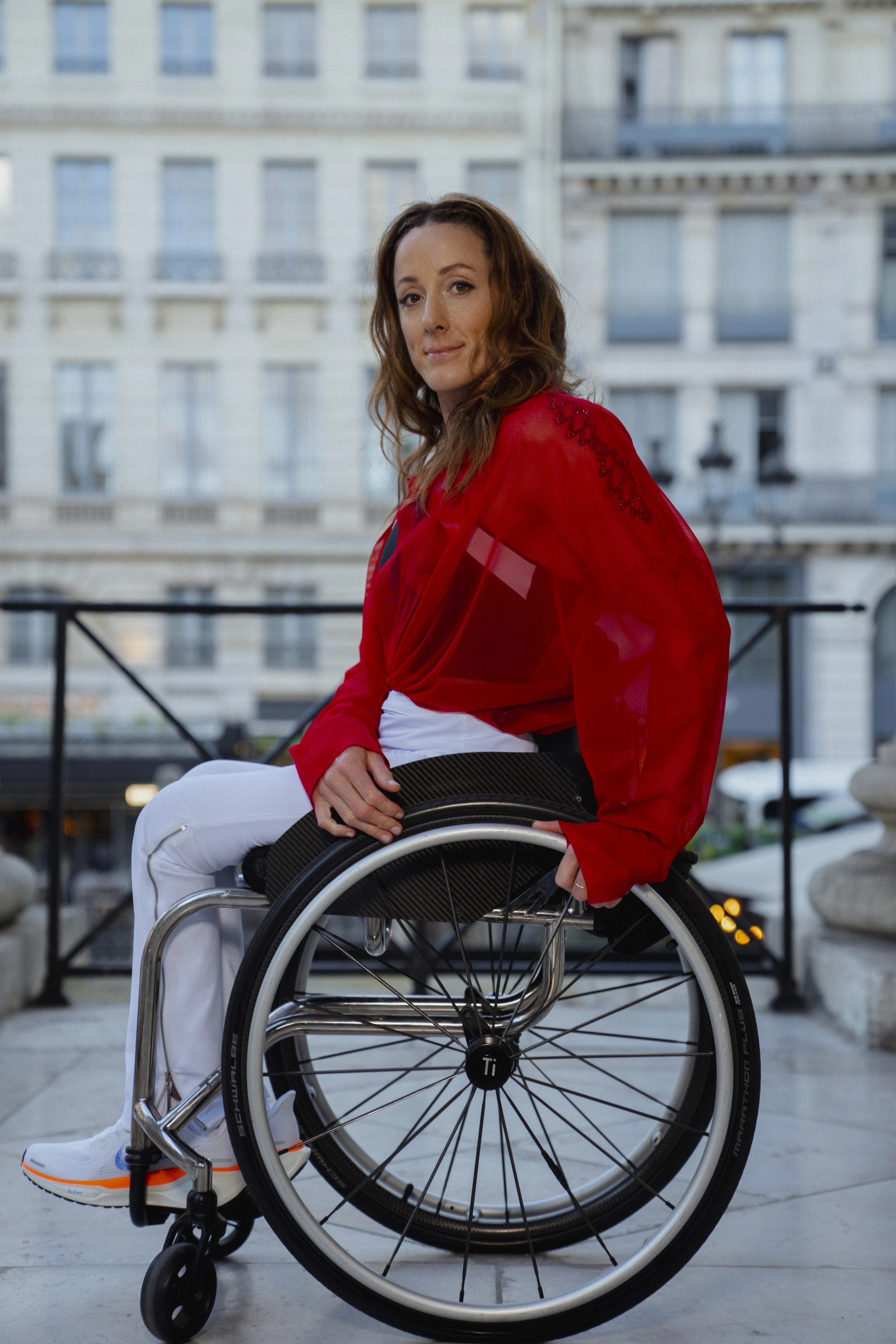 Fully body portrait of Tatyana McFadden sitting in her wheelchair on a marble entryway with European buildings in the background. Tatyana wears a red blouse, white track pants, and Nike Pegasus EasyOn shoes. She smiles towards the camera.