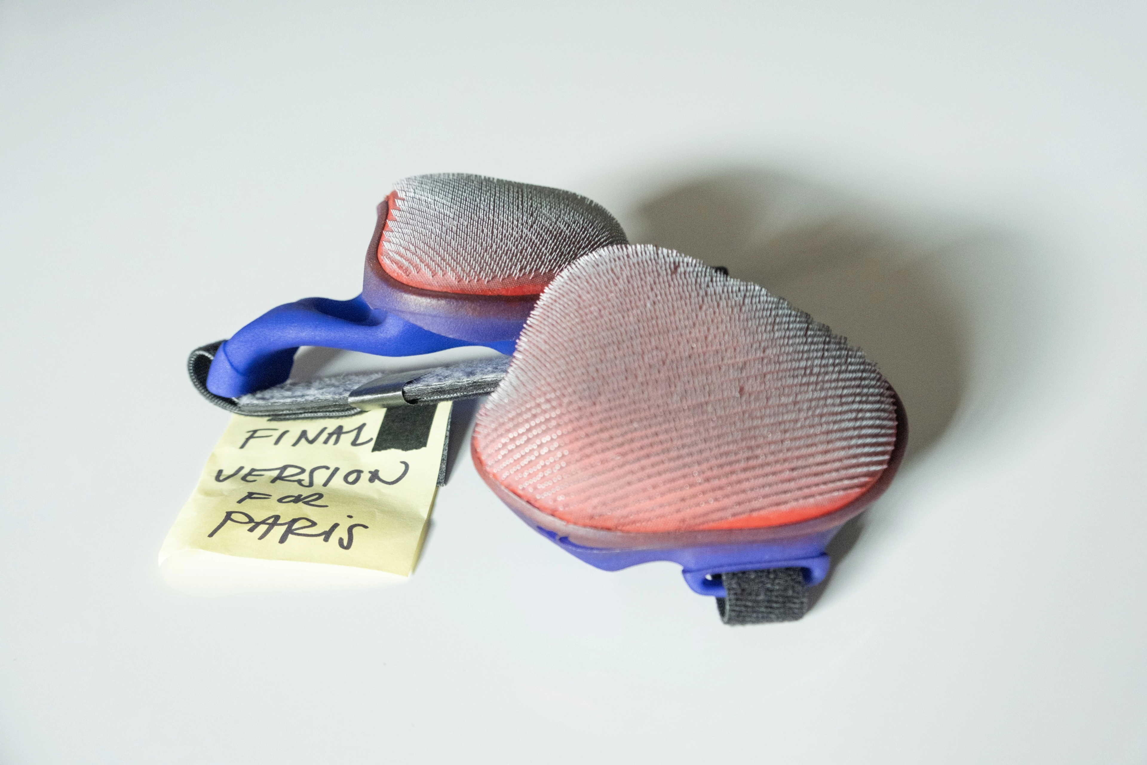 Overhead view of Tatyana McFadden's newly designed Nike push gloves. A pair of blue and red gloves sits on a white surface, alongside a yellow post-it note that reads, "Final version for Paris."