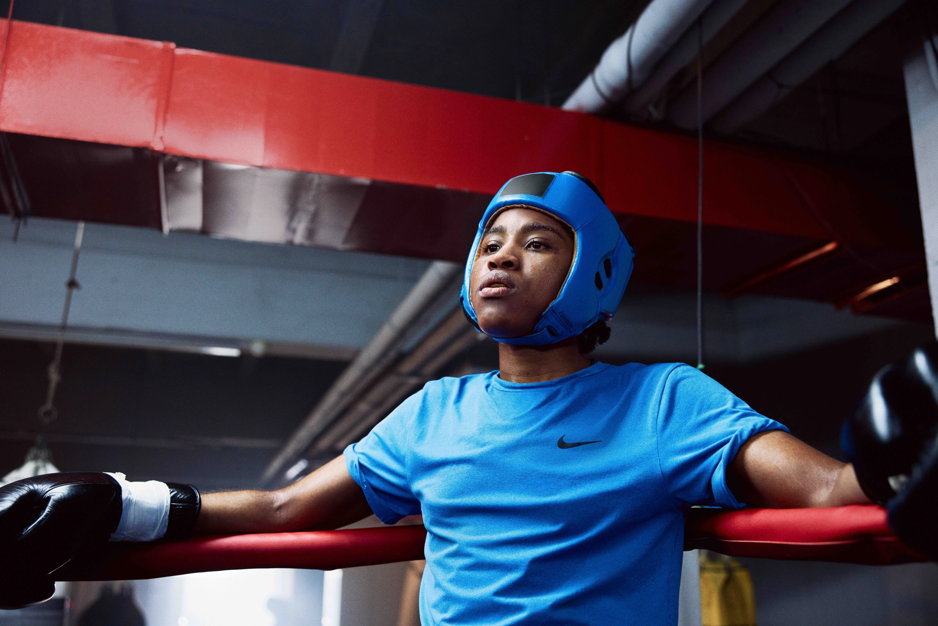 Cindy Ngamba resting against the corner of a boxing ring with arms outstretched resting on the ring ropes and wearing a blue nike shirt, black boxing gloves, and blue protective helmet.