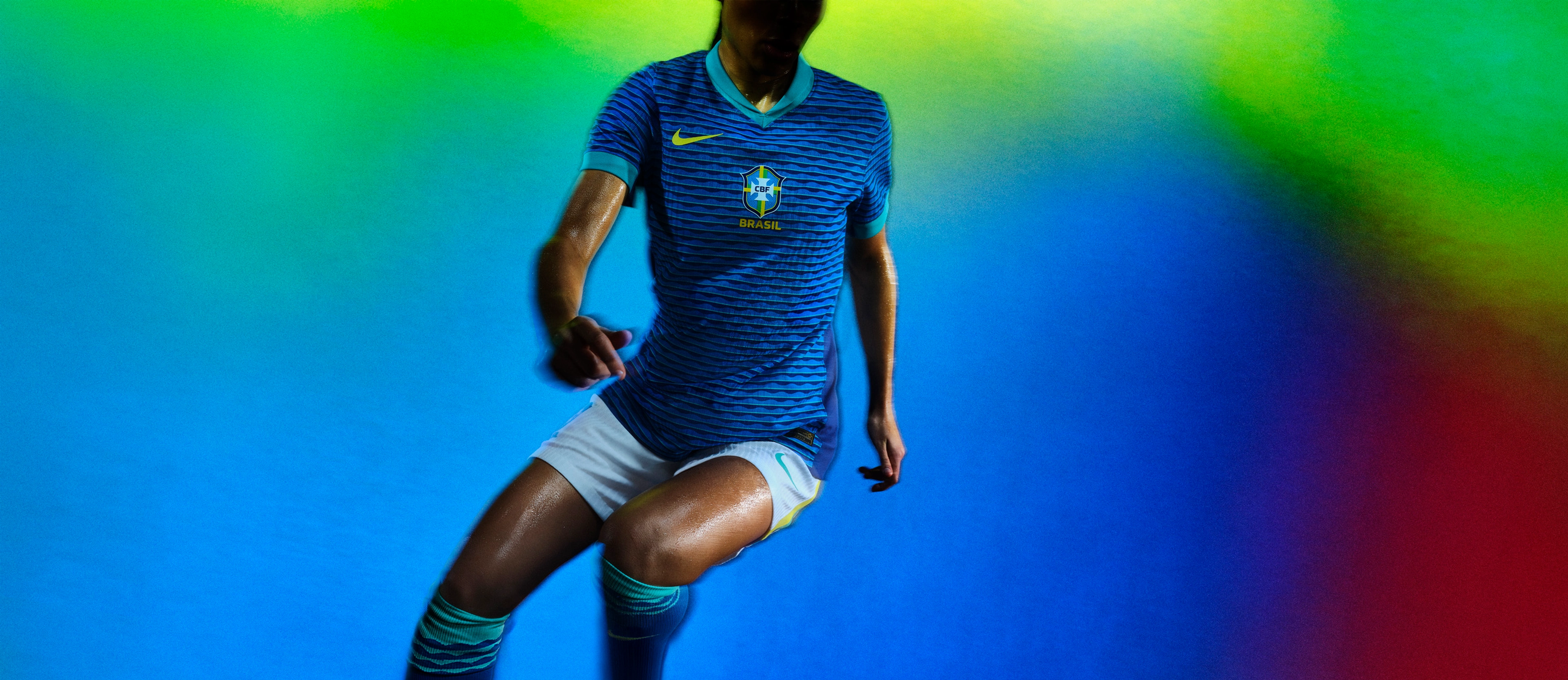 An athlete in a pivoting pose wearing a blue soccer jersey with teal trim on the cuffs and collar, as well as white shorts with a yellow stripe on the side, each with a Nike swoosh and team logo on the chest and legs.