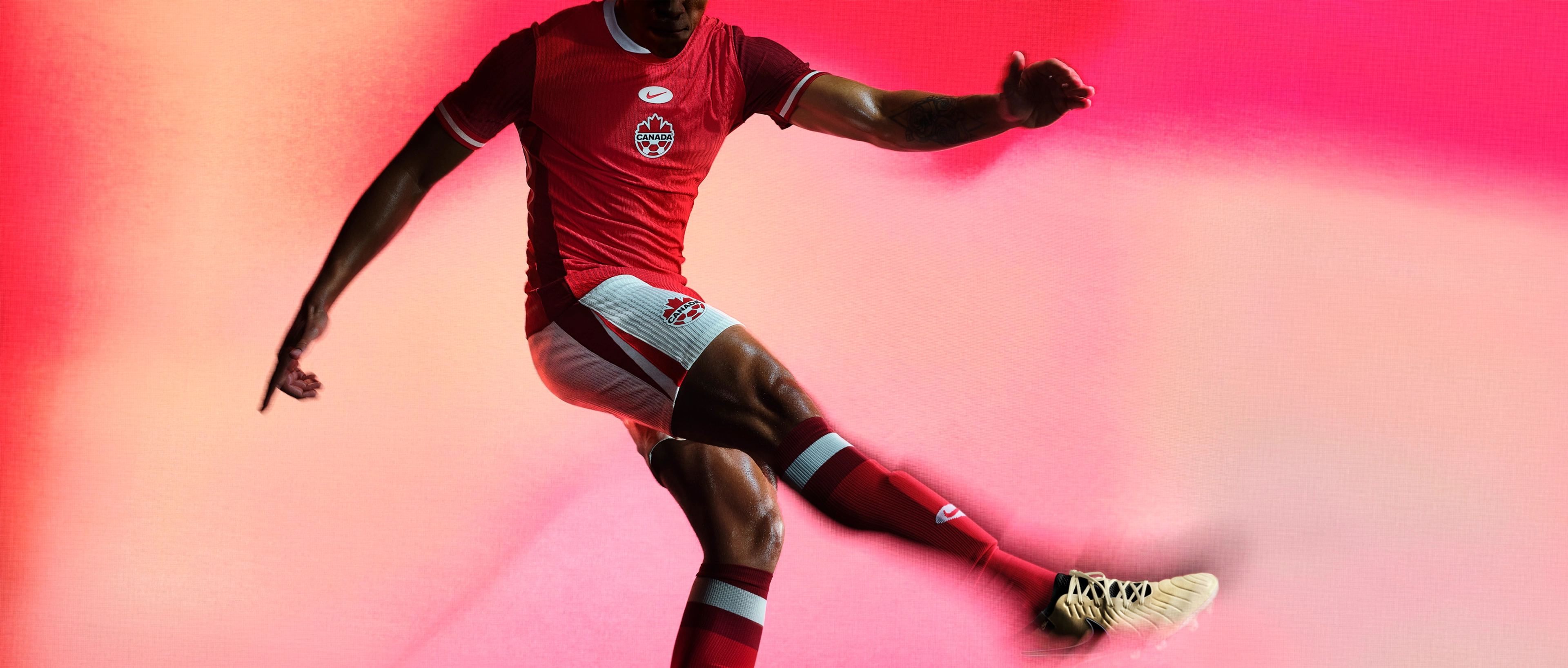 Athlete in a kicking pose wearing a red soccer jersey with red and white striped trim on the collar and cuffs, as well as white shorts with a red and white stripe on the side, each with a Nike swoosh and team logo on the chest and legs.