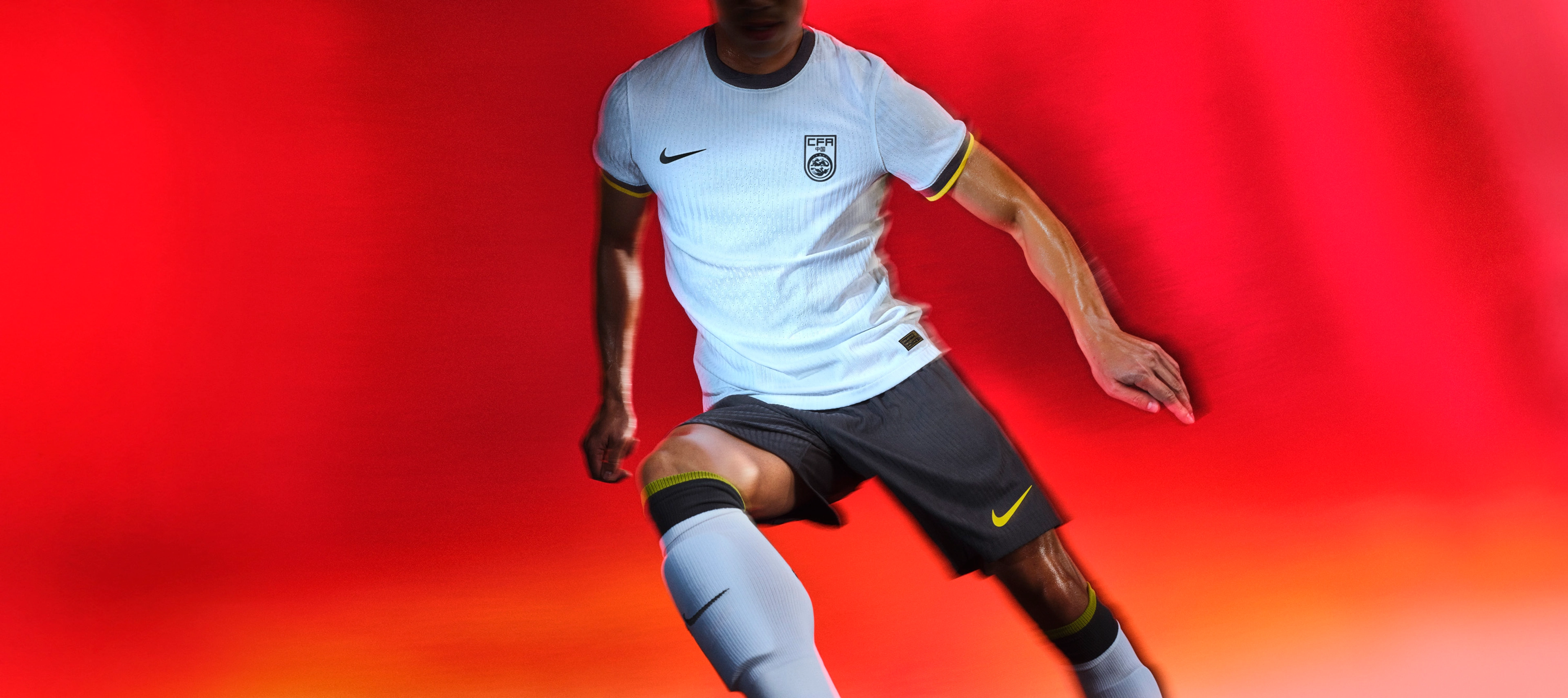 Athlete in a running pose wearing a white soccer jersey with black and yellow striped trim on the cuffs and a black collar, as well as black shorts, each with a Nike swoosh and team logo on the chest and legs. They are also wearing socks with matching colors and trim.