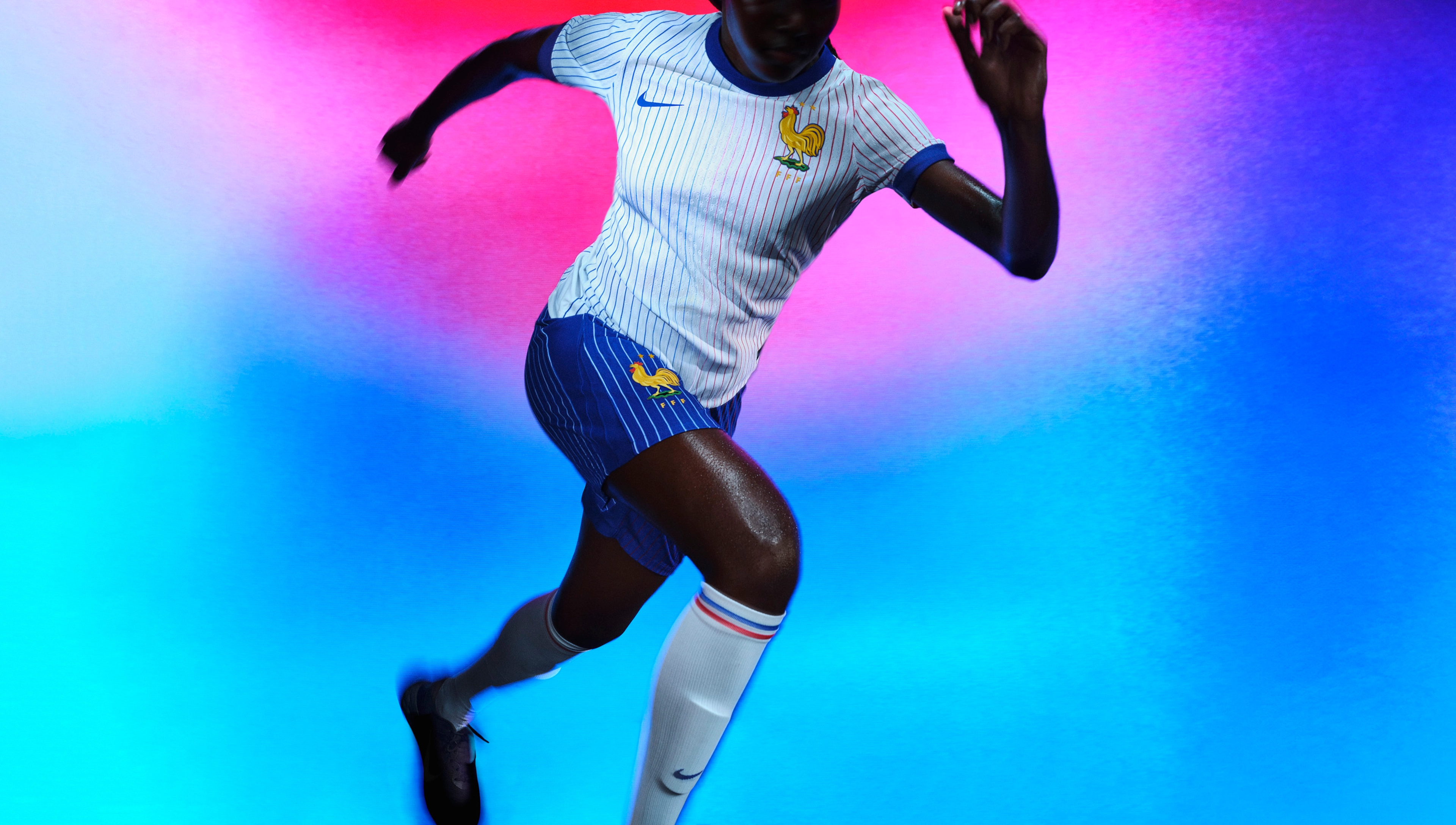 Athlete in a sprinting pose wearing a white soccer jersey with red and blue pinstripes and a blue collar and cuffs, as well as cobalt blue shorts with white pinstripes, each with a Nike swoosh and team logo on the chest and legs.