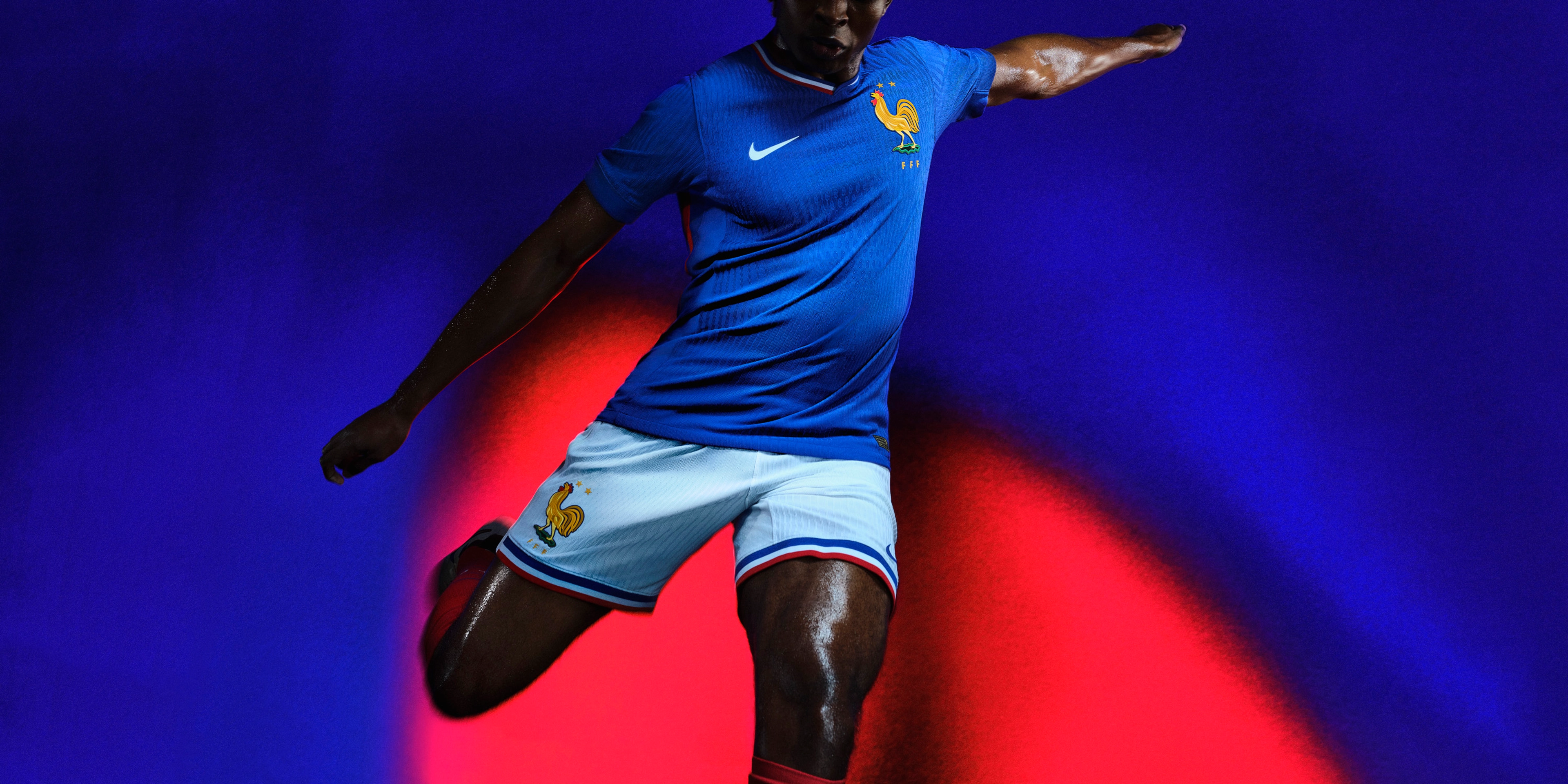 Athlete in a mid-kicking pose wearing a cobalt blue jersey with a red, white, and blue collar, as well as white shorts with red, white, and blue trim, each with a Nike swoosh and team logo on the chest and legs.