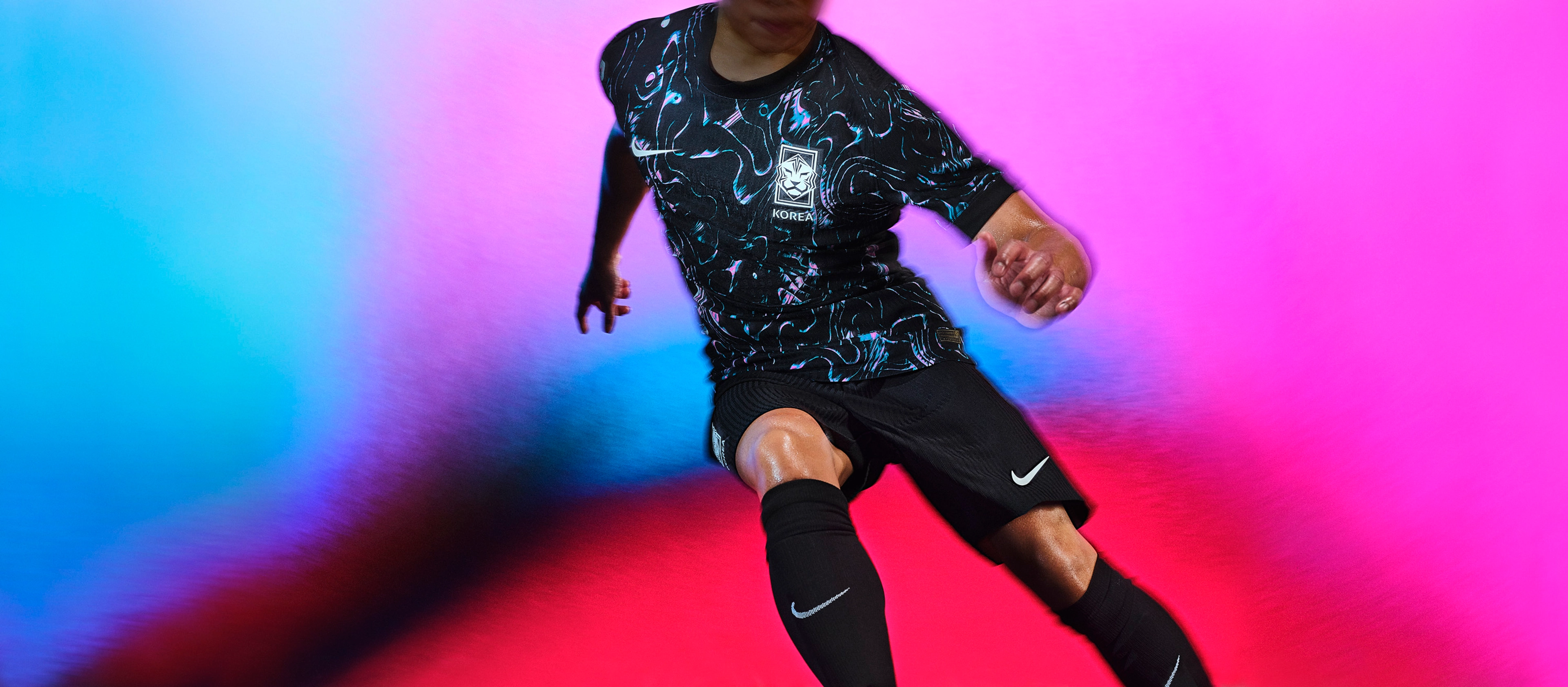 Athlete in a running pose wearing a black soccer jersey with pearlescent pink and blue line patterns, as well as black shorts, each with a Nike swoosh and team on the chest and legs. They are also wearing matching black socks.